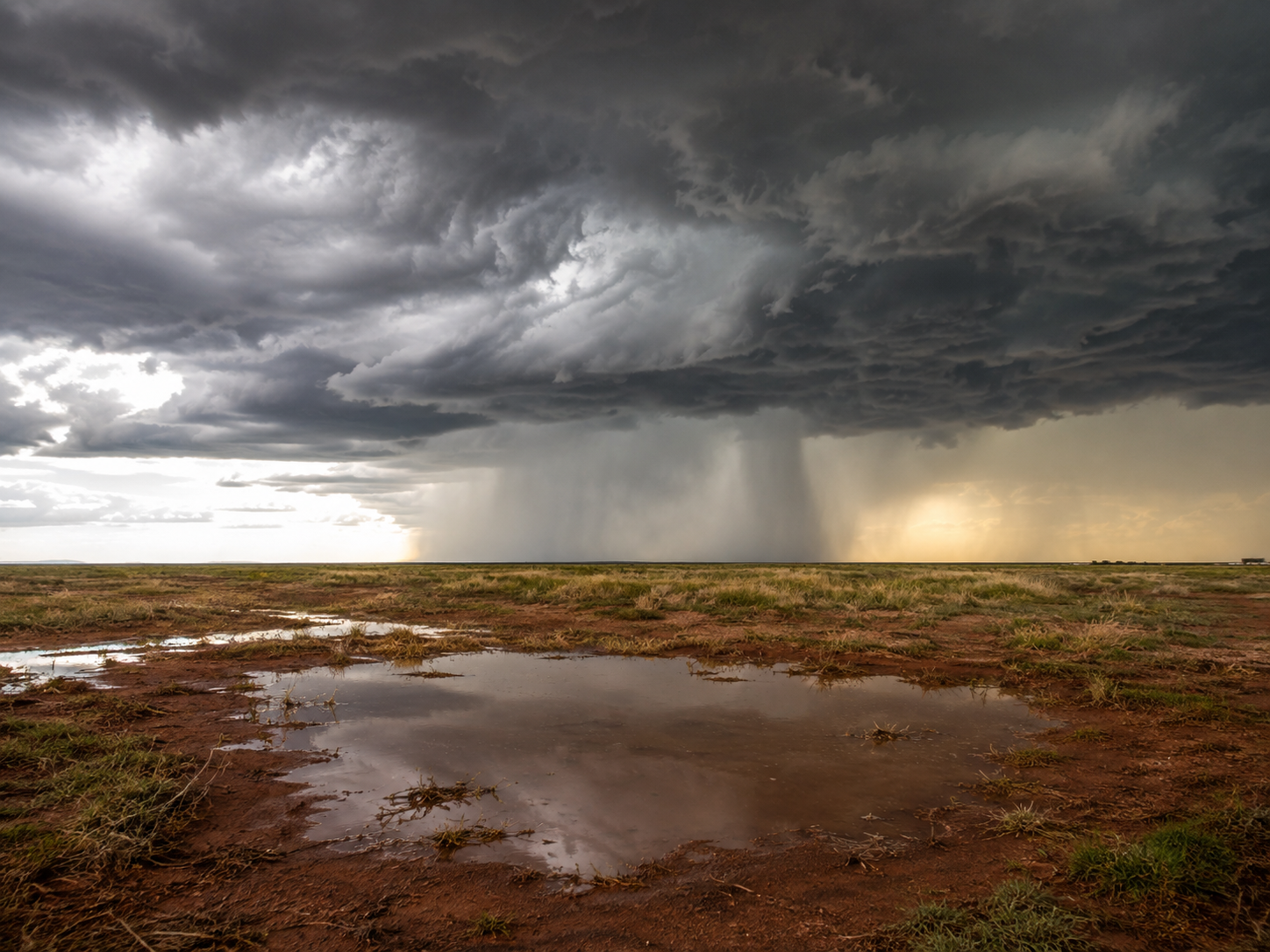 Dramatic storm over red dirt plains with rain curtain, dark clouds, and a muddy puddle reflecting the turbulent sky.