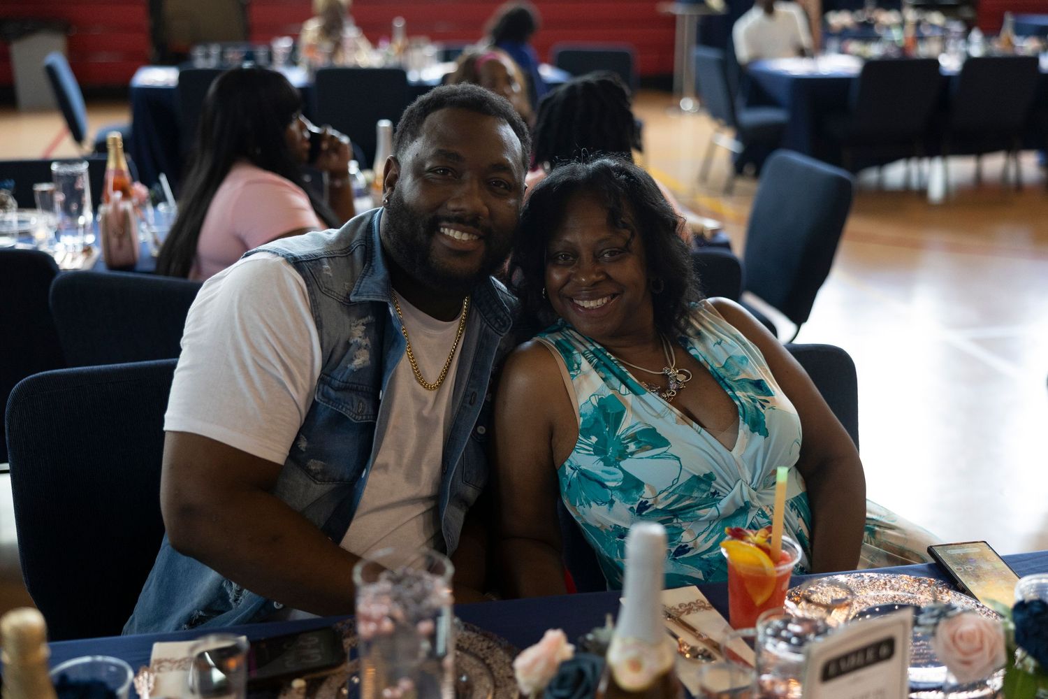 Two smiling people pose together at a banquet event, seated at a decorated table in a large hall.