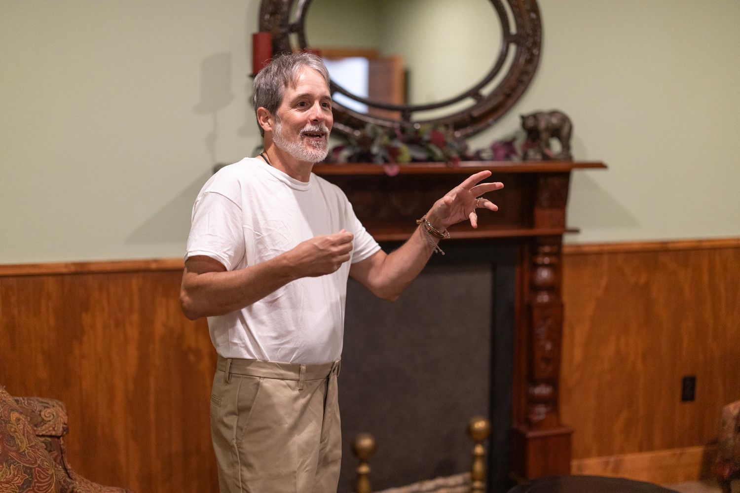 Smiling older man with gray beard wearing apron gesturing while standing near decorative fireplace indoors.
