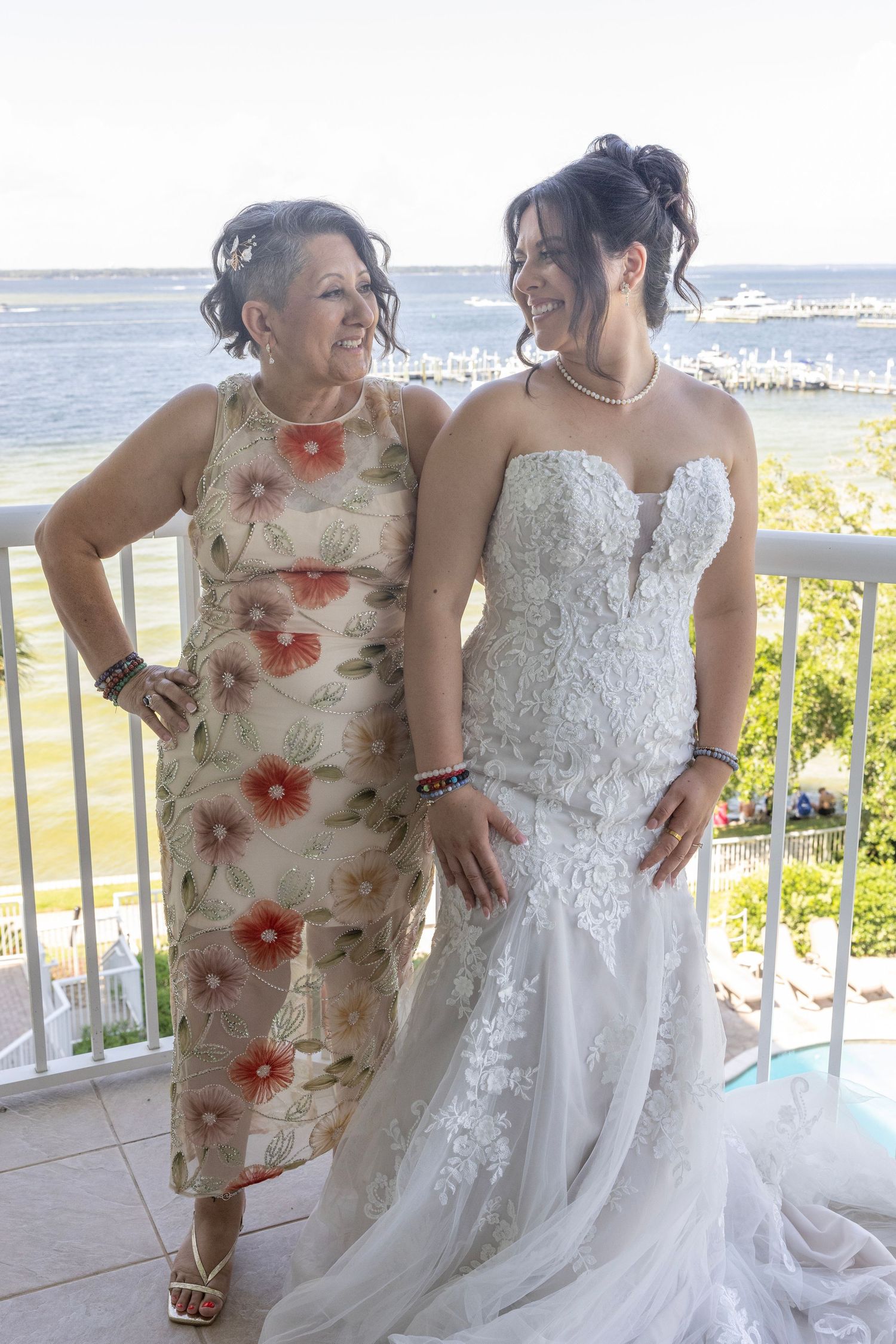 A bride in a white lace gown smiles with an older woman in a floral dress on a waterfront balcony.