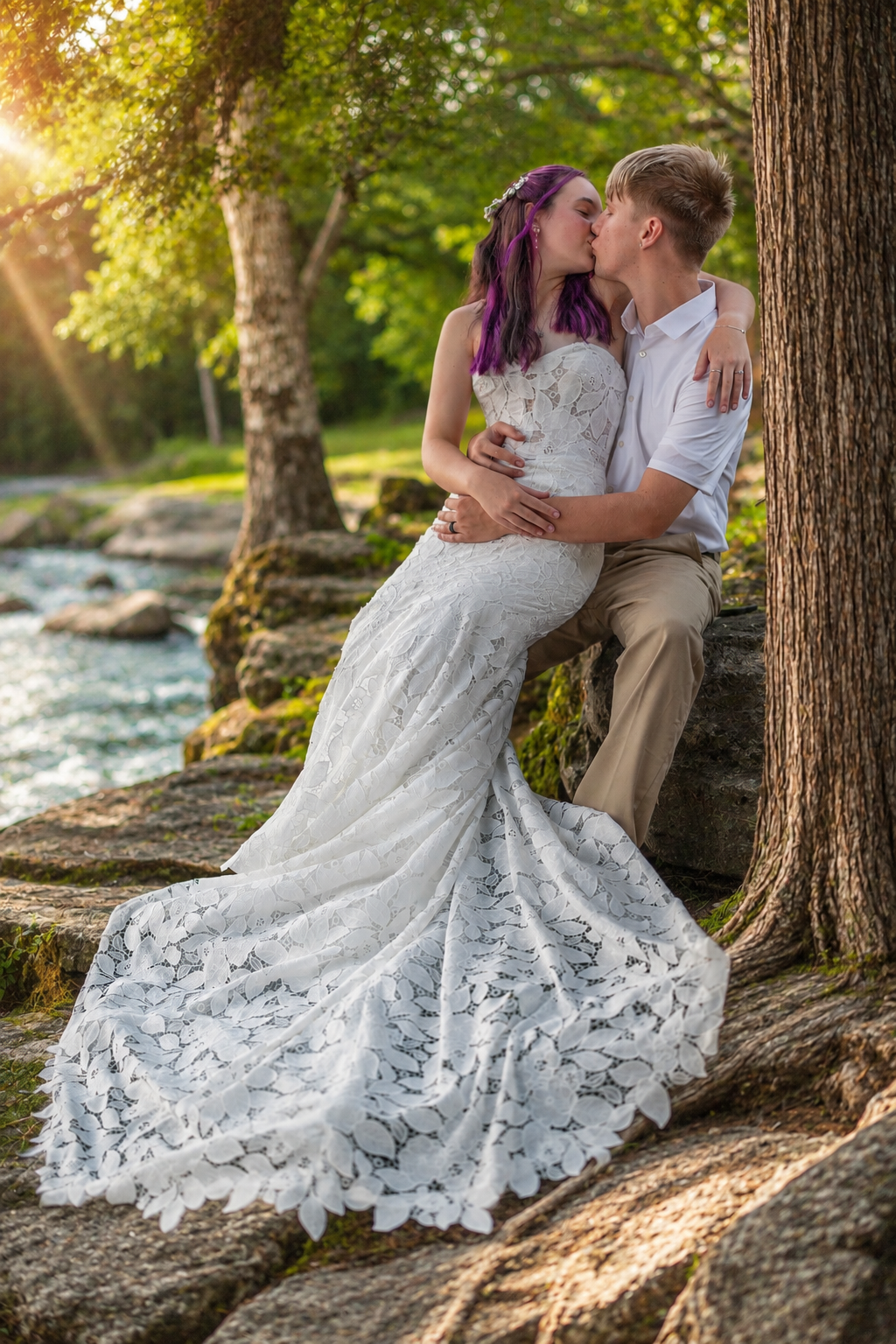 Bride in white lace gown and groom share a kiss by a sunlit river, surrounded by lush green trees.