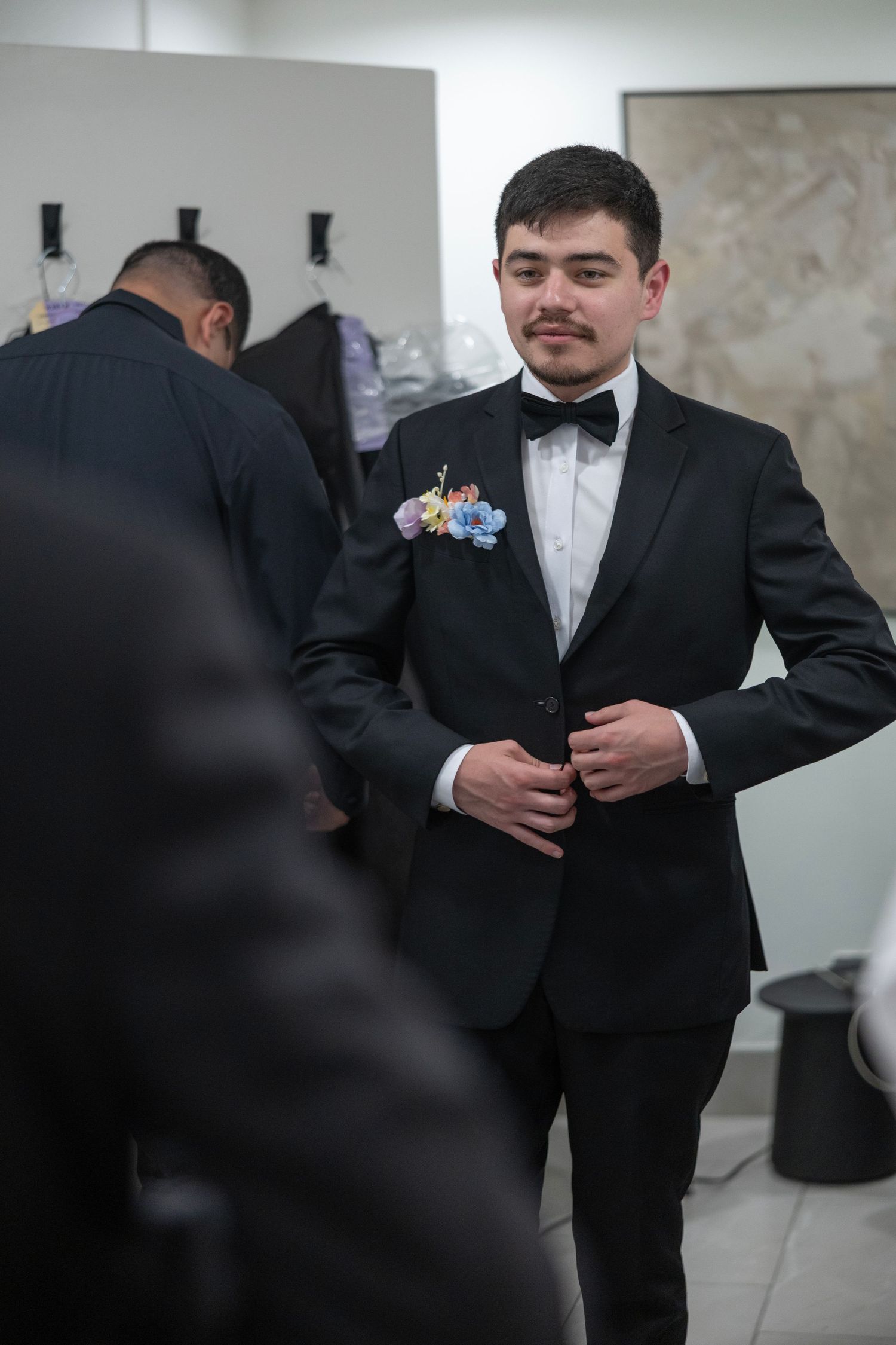 Young man in black tuxedo with floral boutonniere buttoning jacket in formal dressing room.