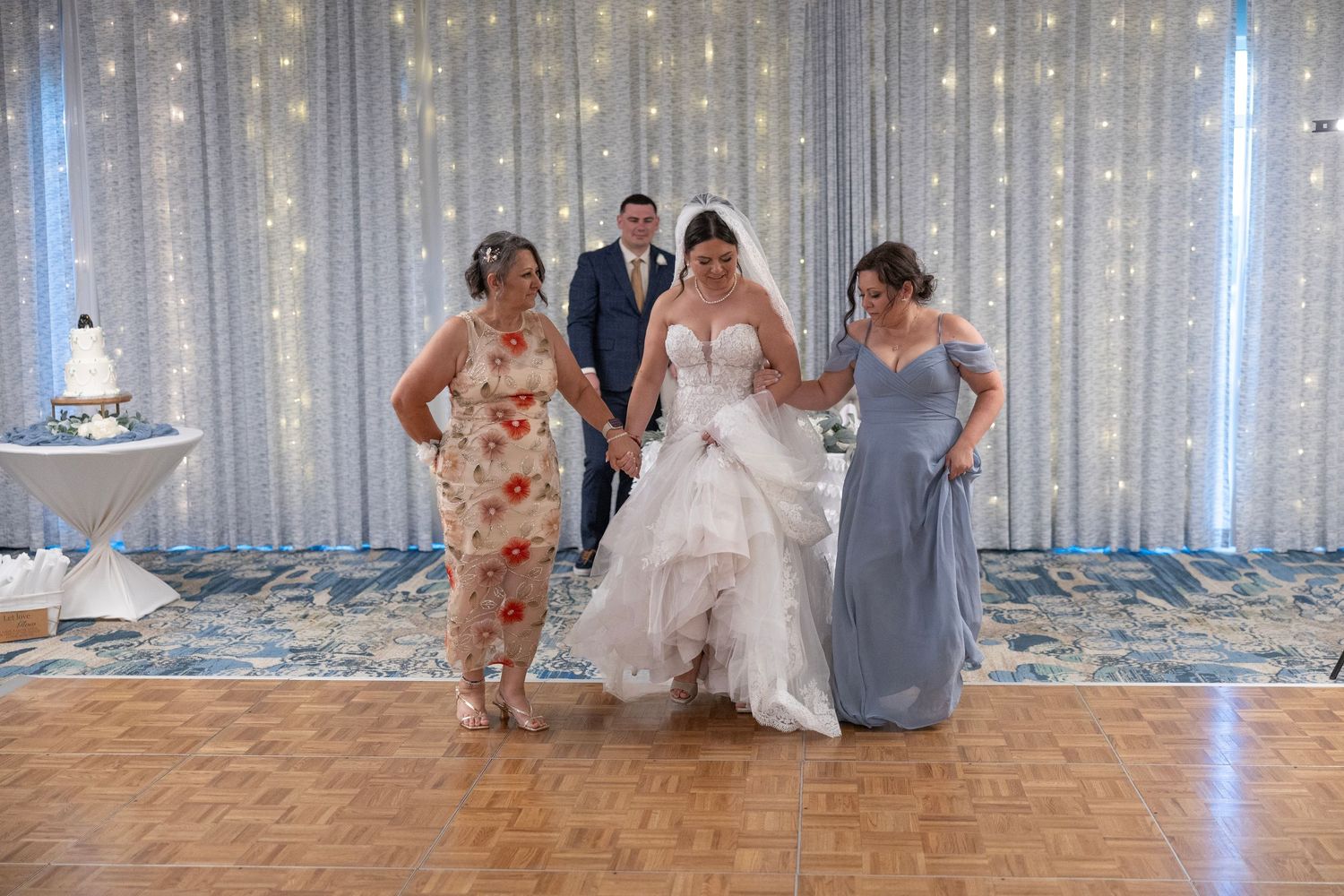 Bride in white gown assisted by guests on dance floor at elegant wedding reception with fairy light backdrop.