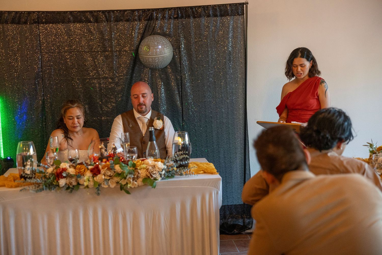 Woman in red dress giving a speech at a wedding reception dinner while bride and groom sit at the head table.