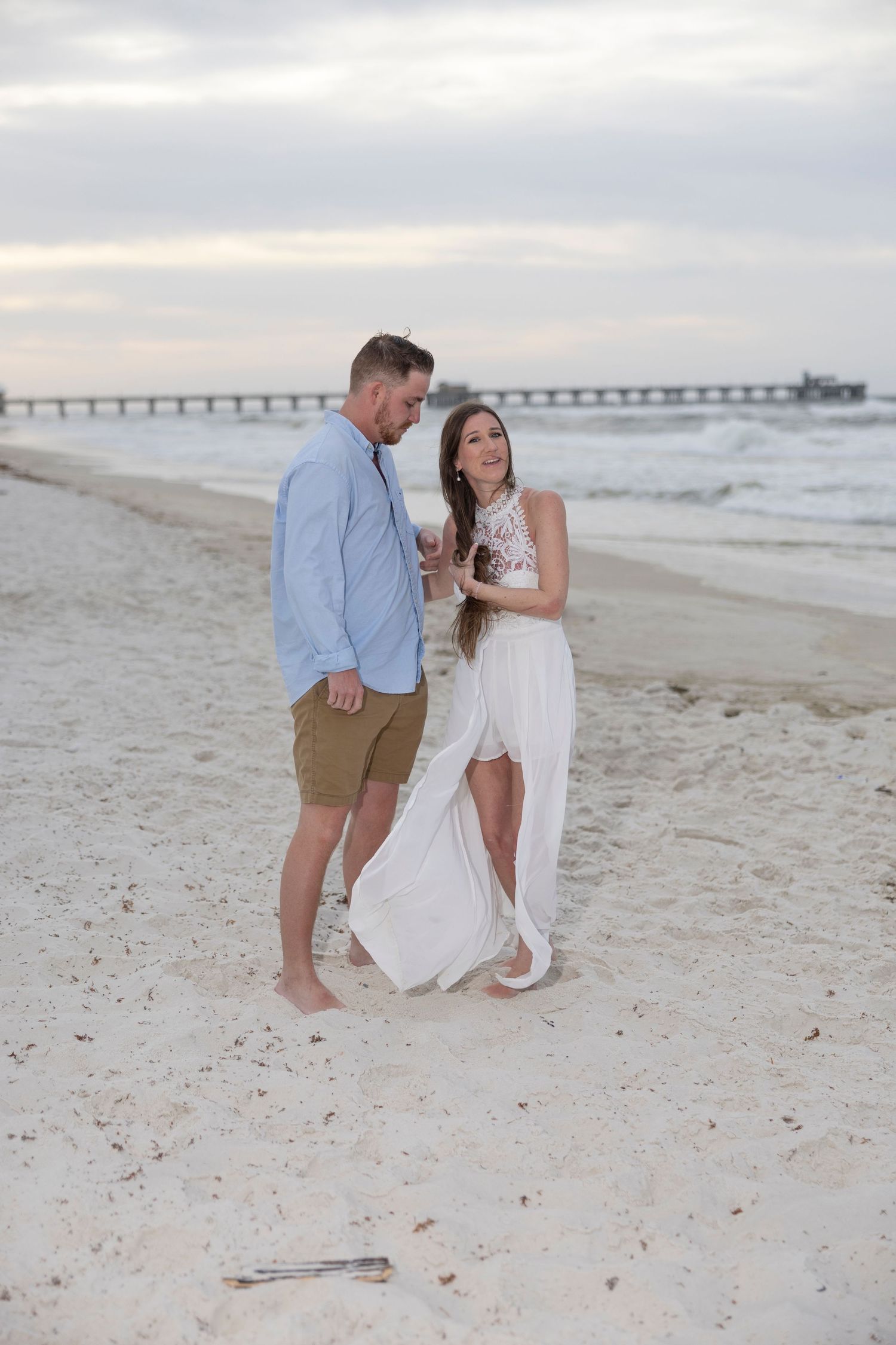 Couple in elegant beach attire sharing a romantic moment on sandy shore at sunset with pier in background.
