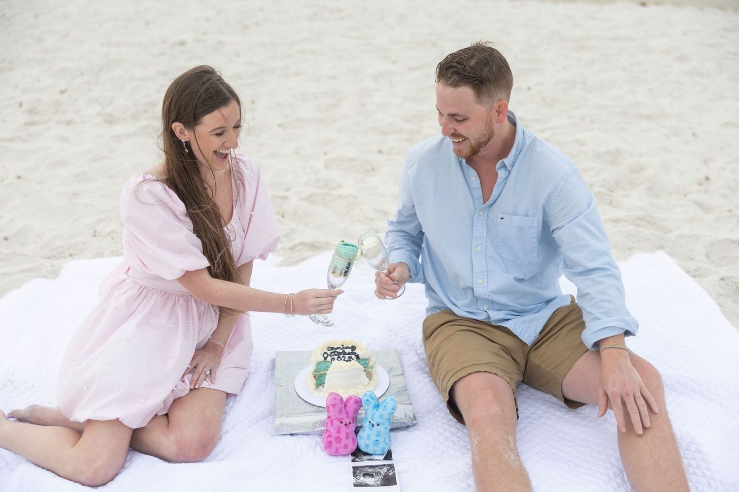 Couple enjoying a romantic beach picnic with cake and drinks on a white blanket on sandy shore.