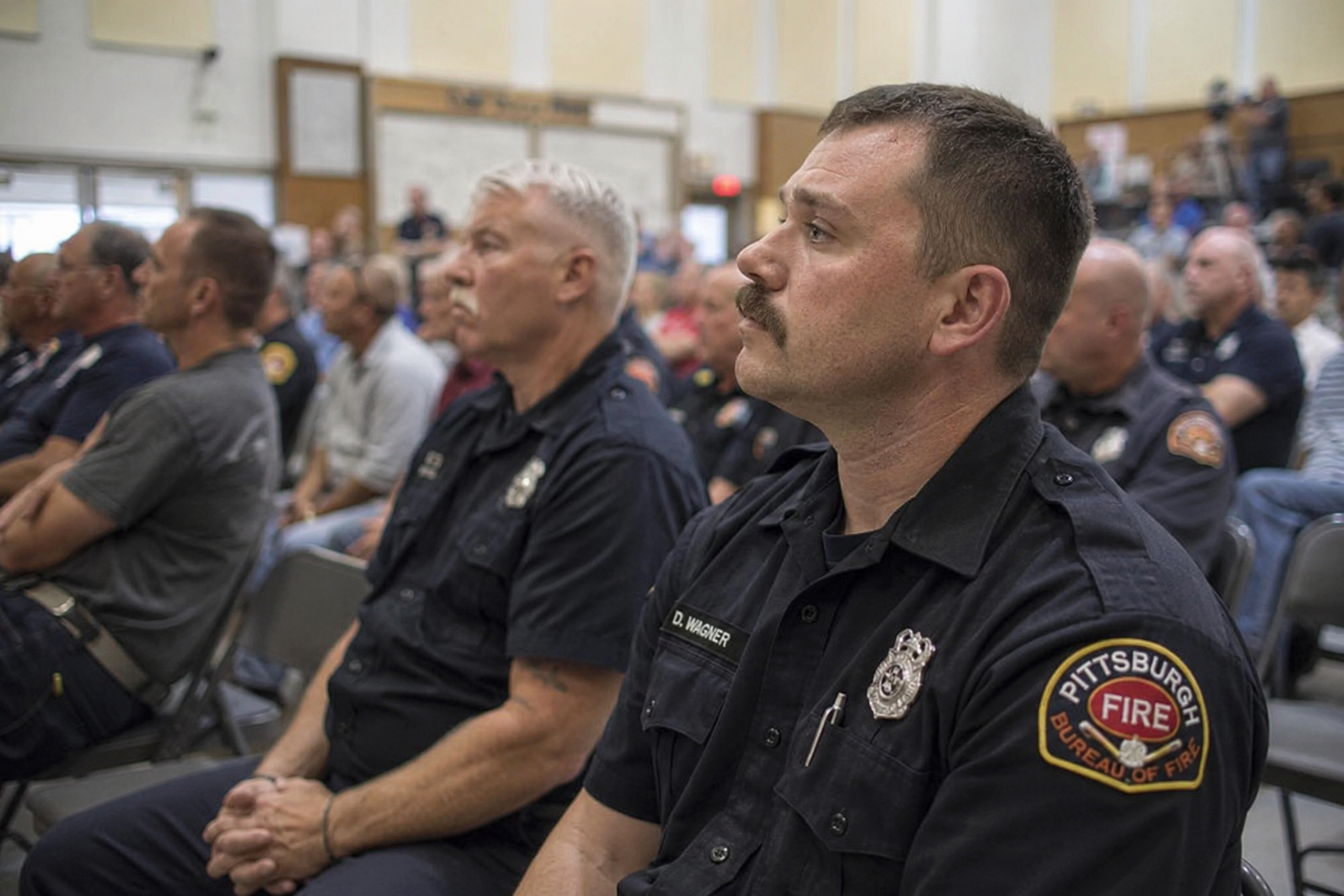Pittsburgh firefighters in uniform sit attentively during an official meeting or ceremony in a large assembly hall.