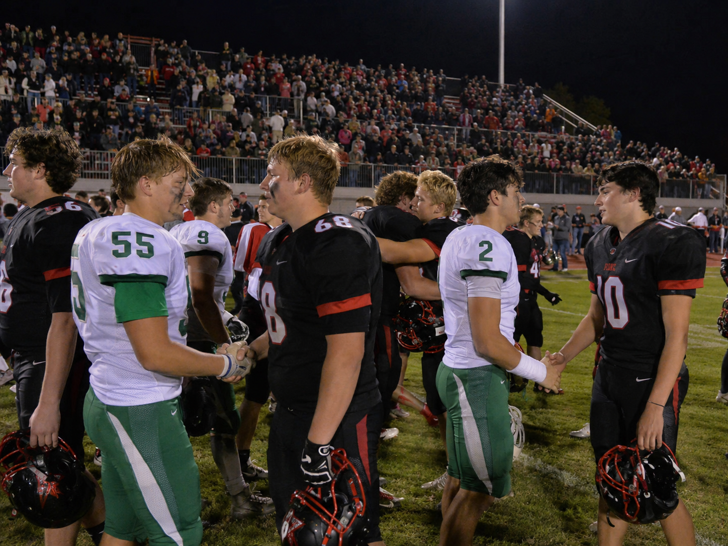 High school football players from opposing teams shake hands on the field after a night game with packed bleachers.