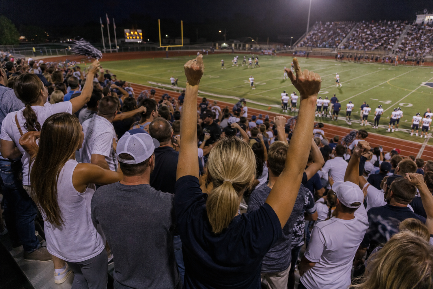 Excited fans cheer with raised arms in packed stadium stands during a night football game.