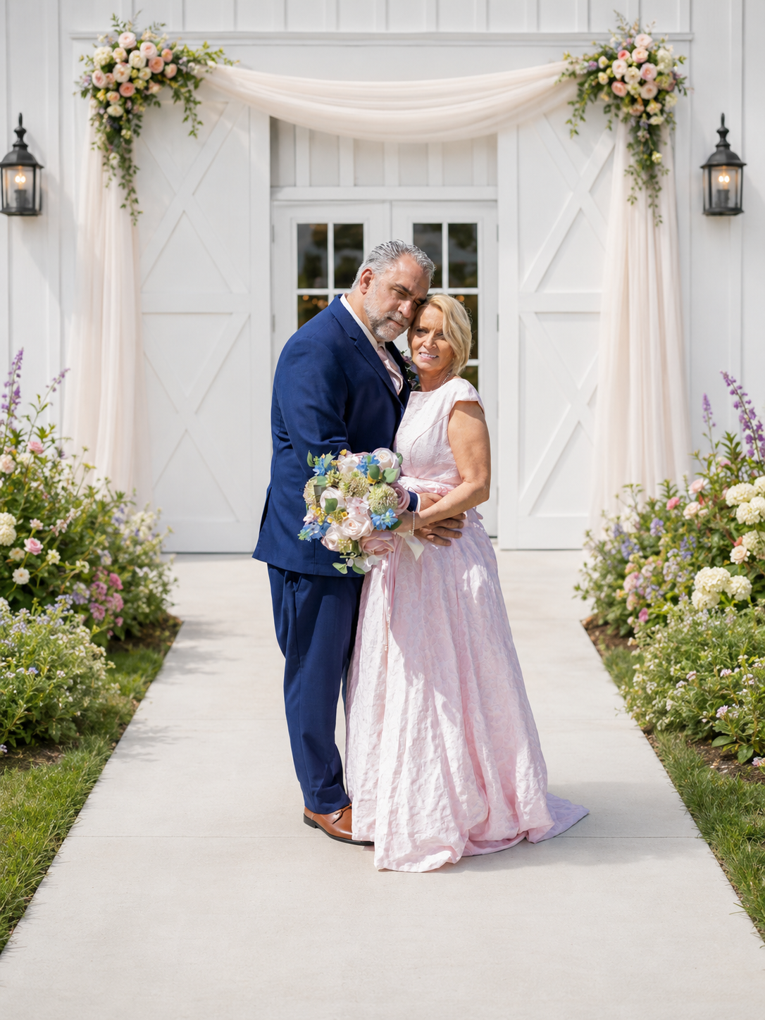 Elegant couple in pink gown and navy suit posing before white barn adorned with floral arrangements.