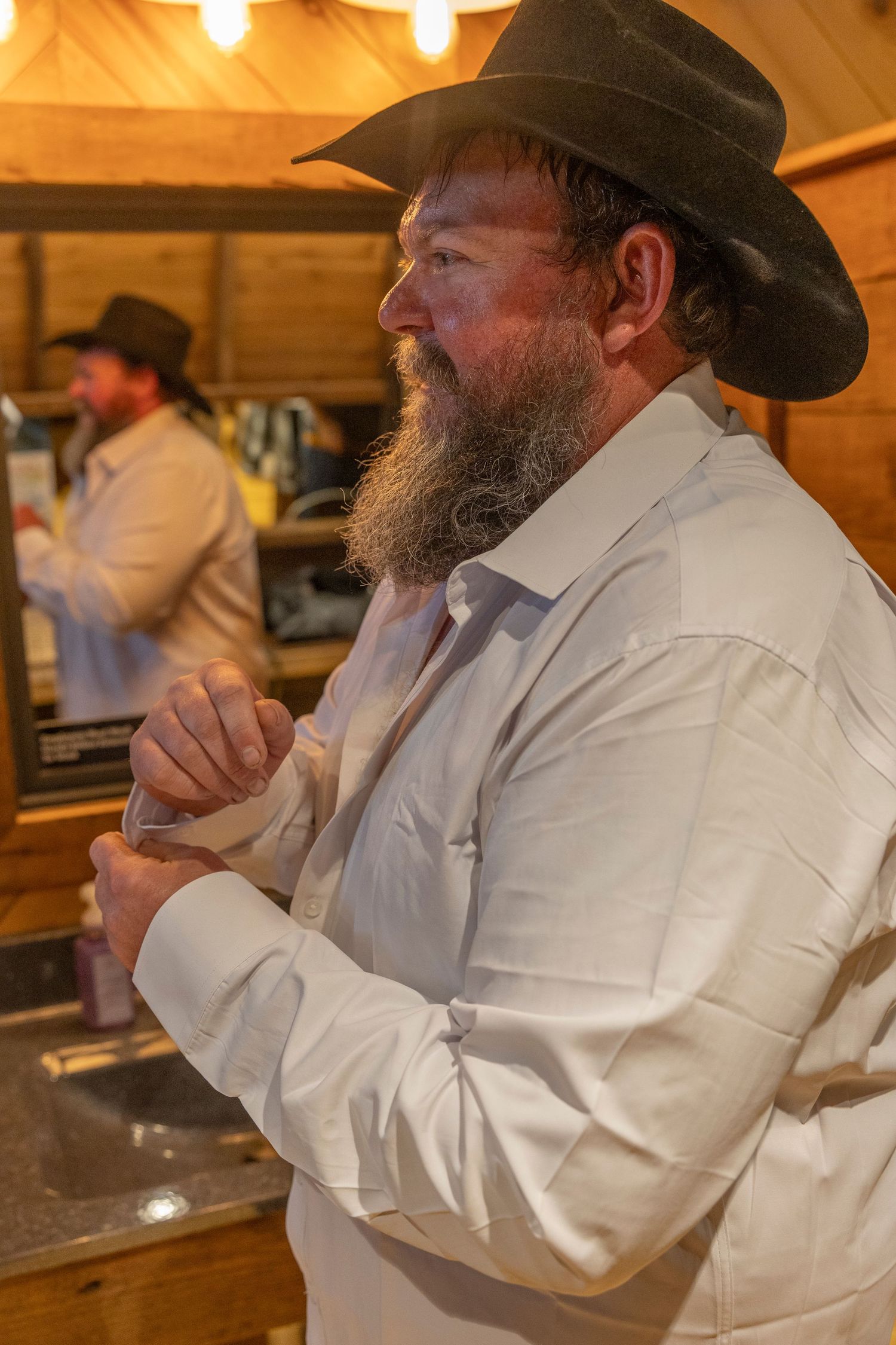 Bearded man in cowboy hat and white shirt stands at a rustic wooden bar, with his reflection visible in a mirror.