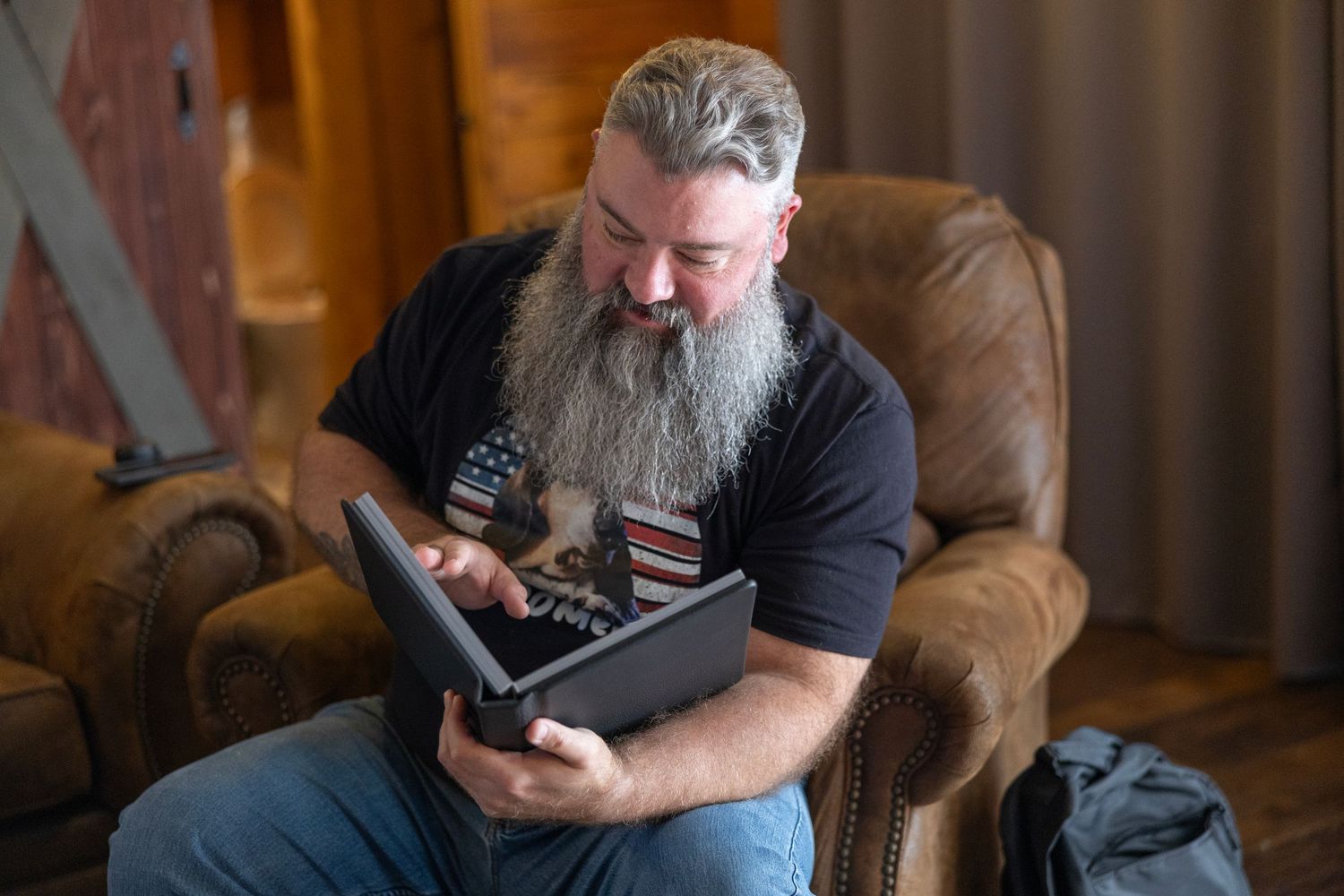 Bearded man with gray hair sits in leather armchair using laptop computer indoors.