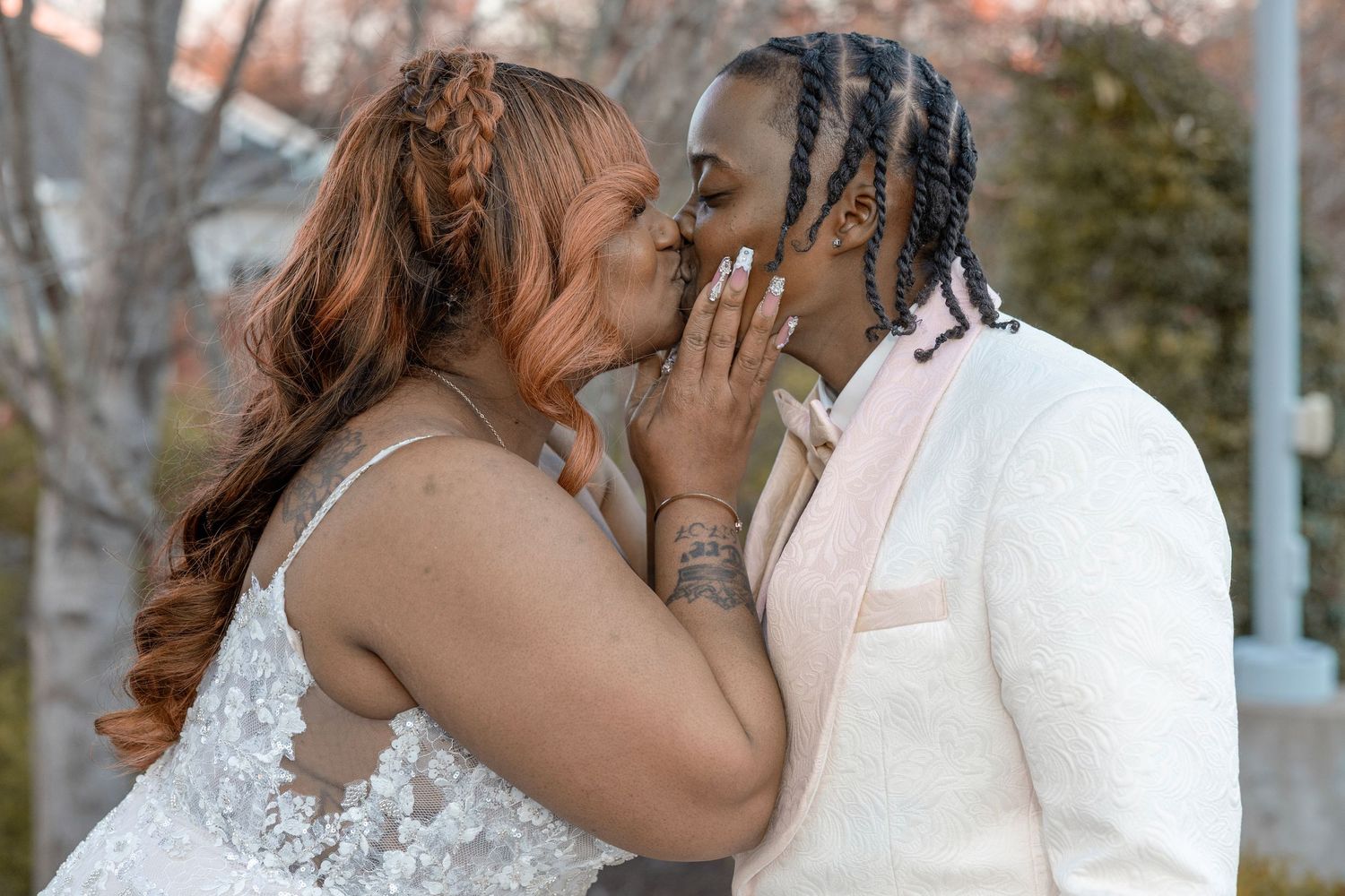 A couple shares a romantic kiss in wedding attire outdoors, bride in lace gown and groom in white suit.