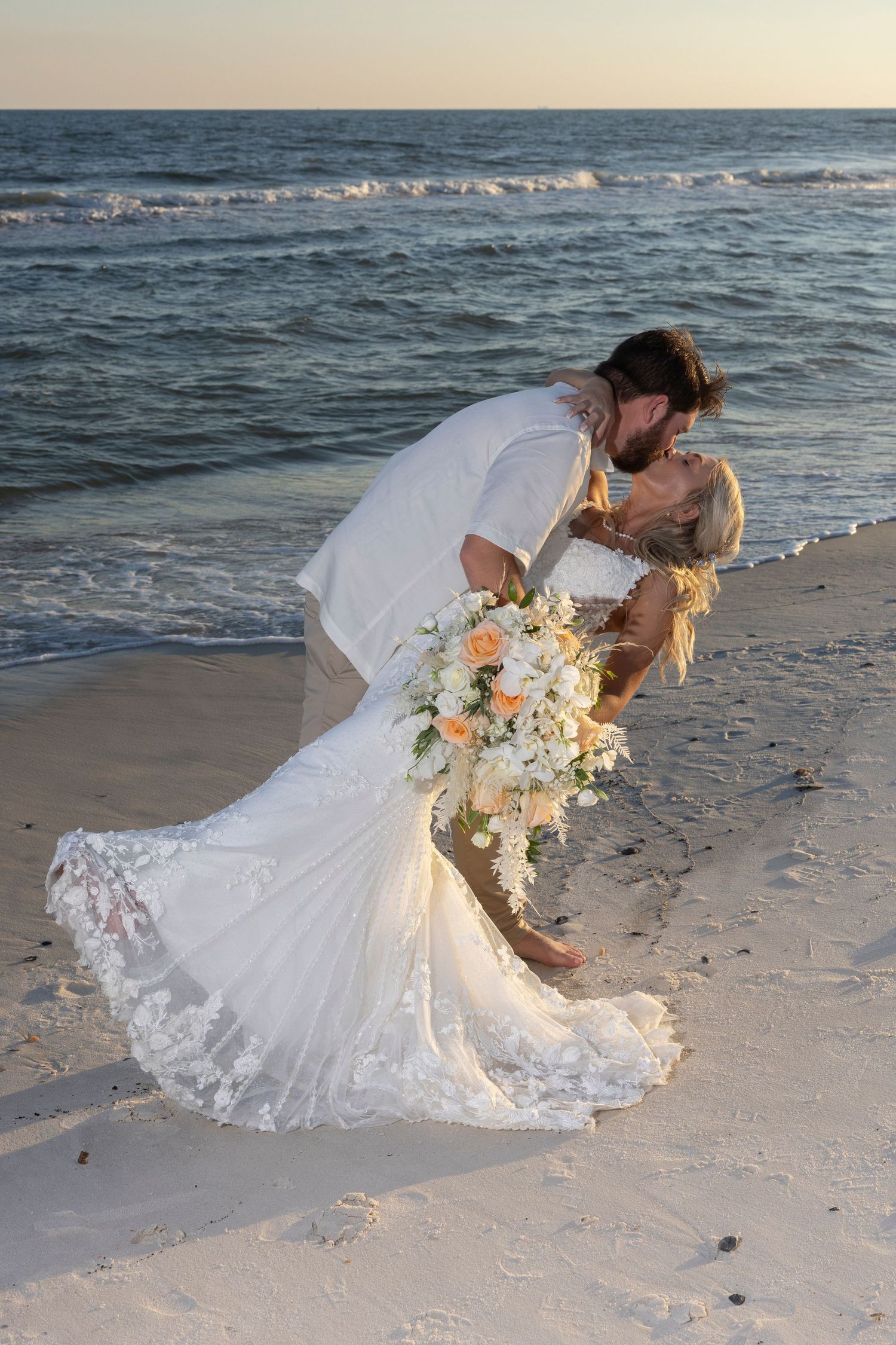A bride in a flowing white lace gown poses gracefully on a sandy beach at sunset with her floral bouquet.