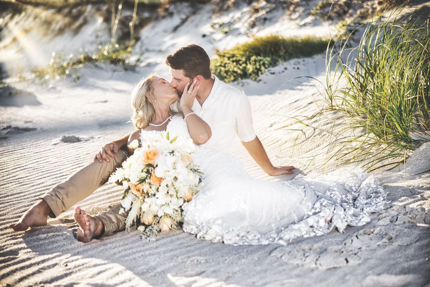 A romantic beach wedding couple sits together in soft sand among coastal grass and dunes at sunset.