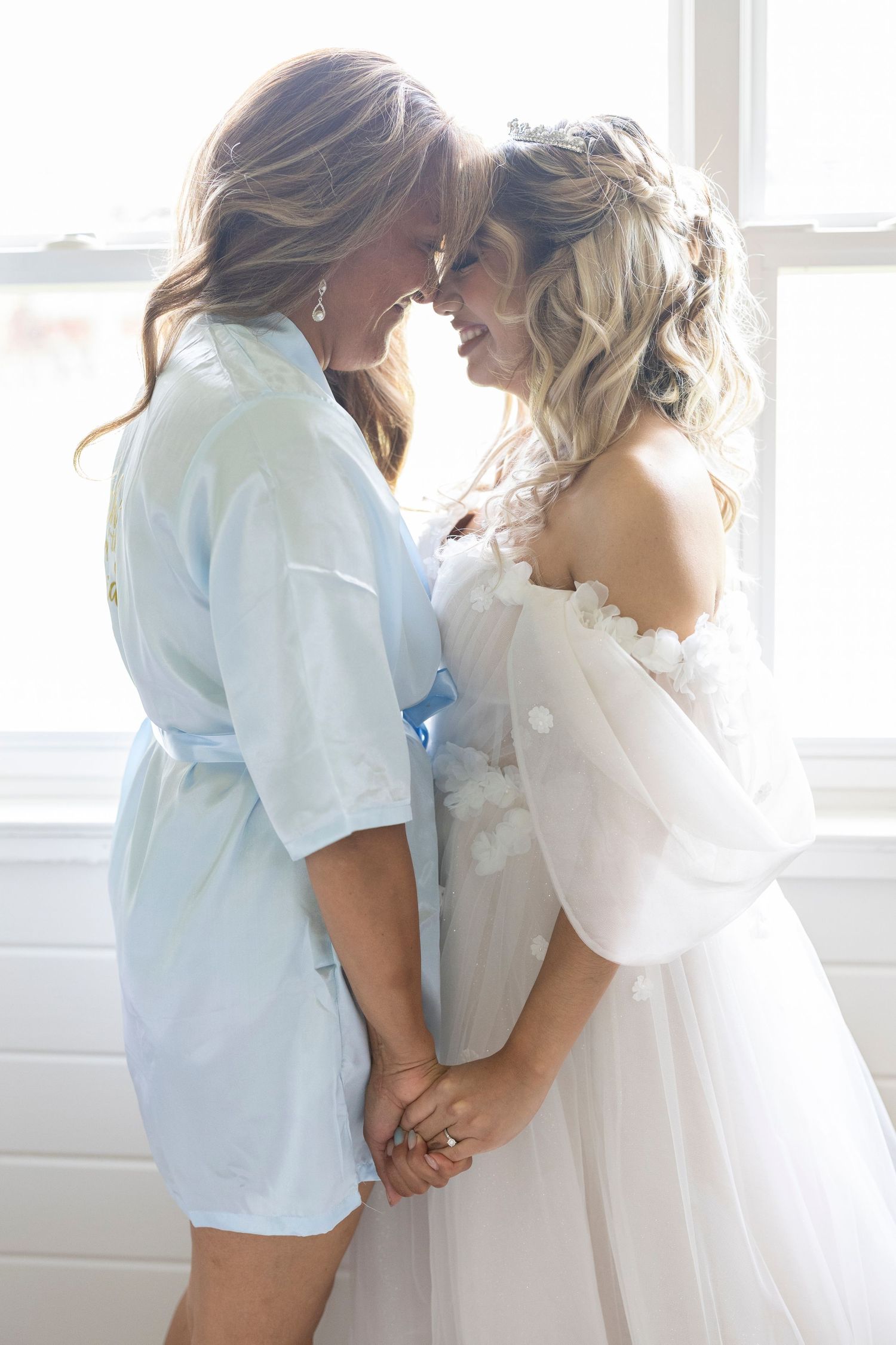 Two people embrace in a romantic moment by a bright window wearing white and light blue clothing.