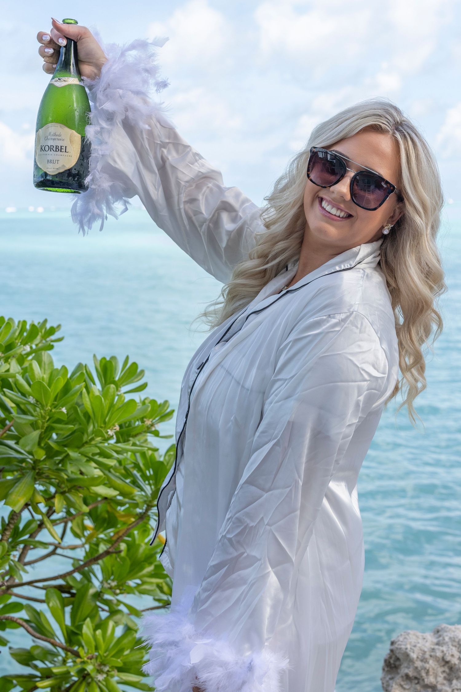 Person in white shirt poses with champagne bottle against turquoise ocean backdrop on sunny tropical day.