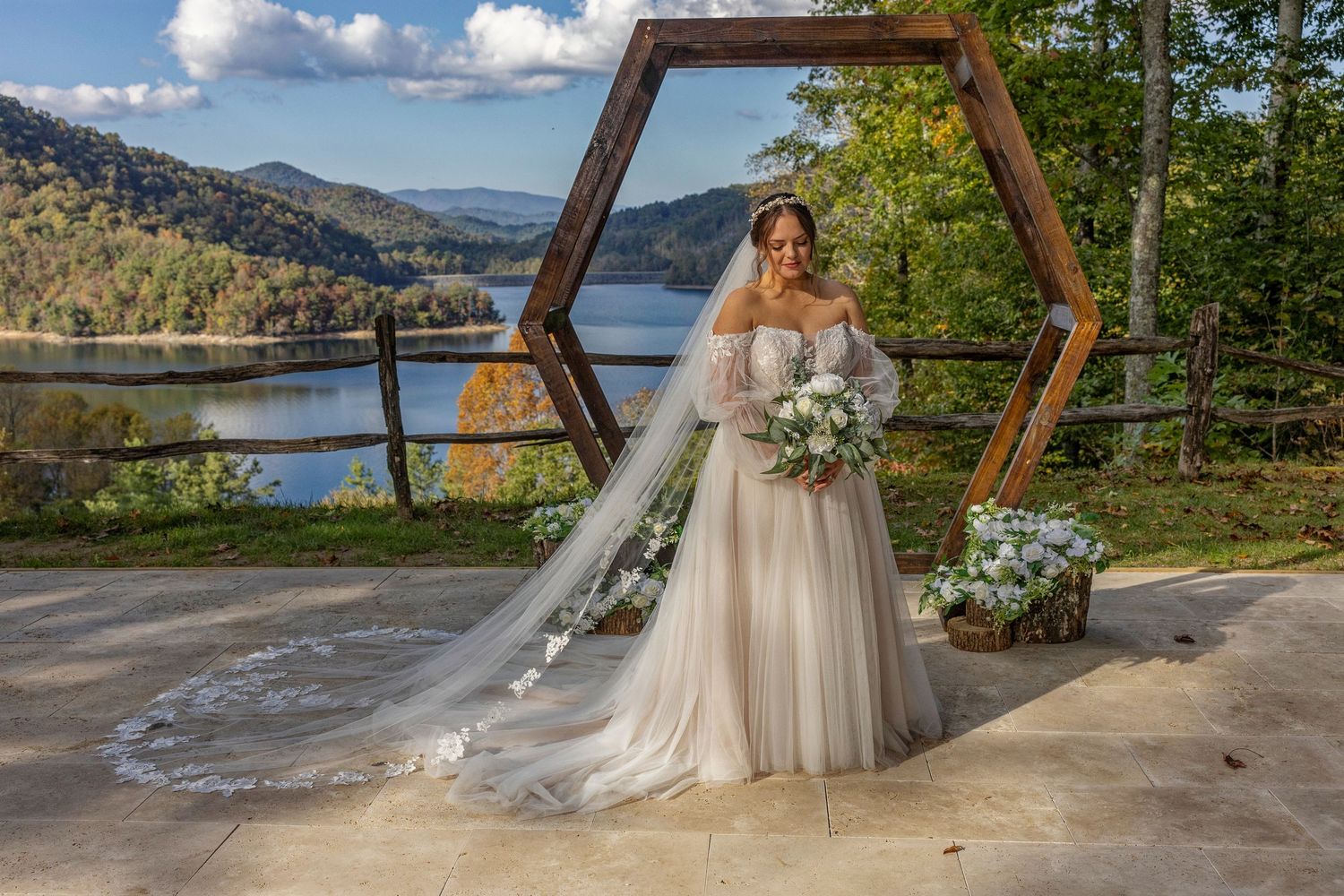 A scenic outdoor wedding ceremony setup overlooking a mountain lake with a hexagonal wooden arch and white floral decor.
