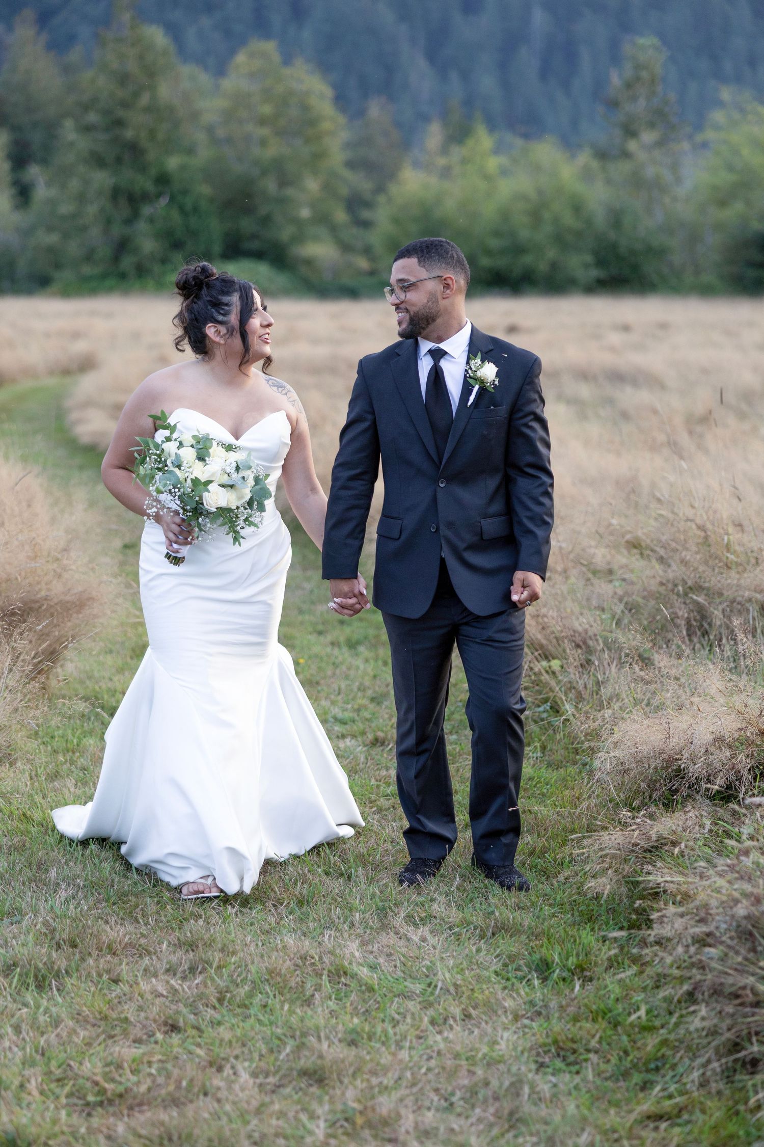 A couple in wedding attire stands together in a scenic field with mountains and trees in the background.