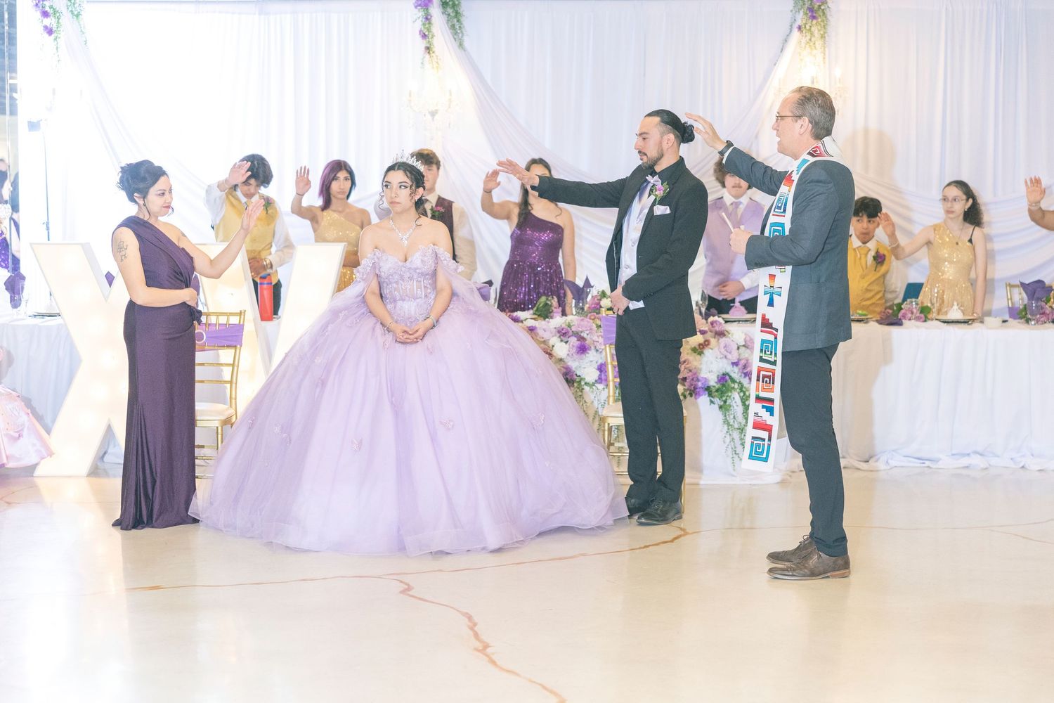 A quinceañera celebration in a banquet hall shows guests gathered around a person in a lavender ball gown.