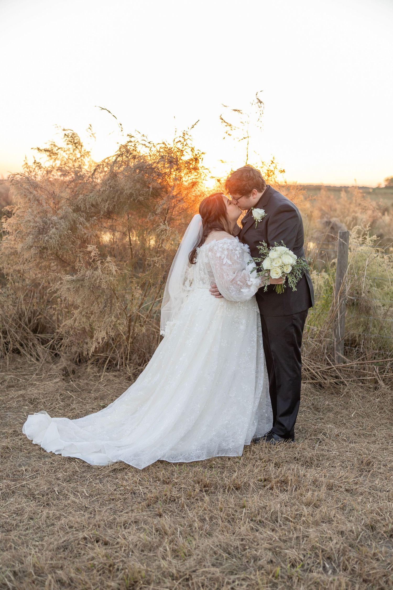 A romantic wedding couple embraces during golden hour in a rustic field with dried grass and winter foliage.