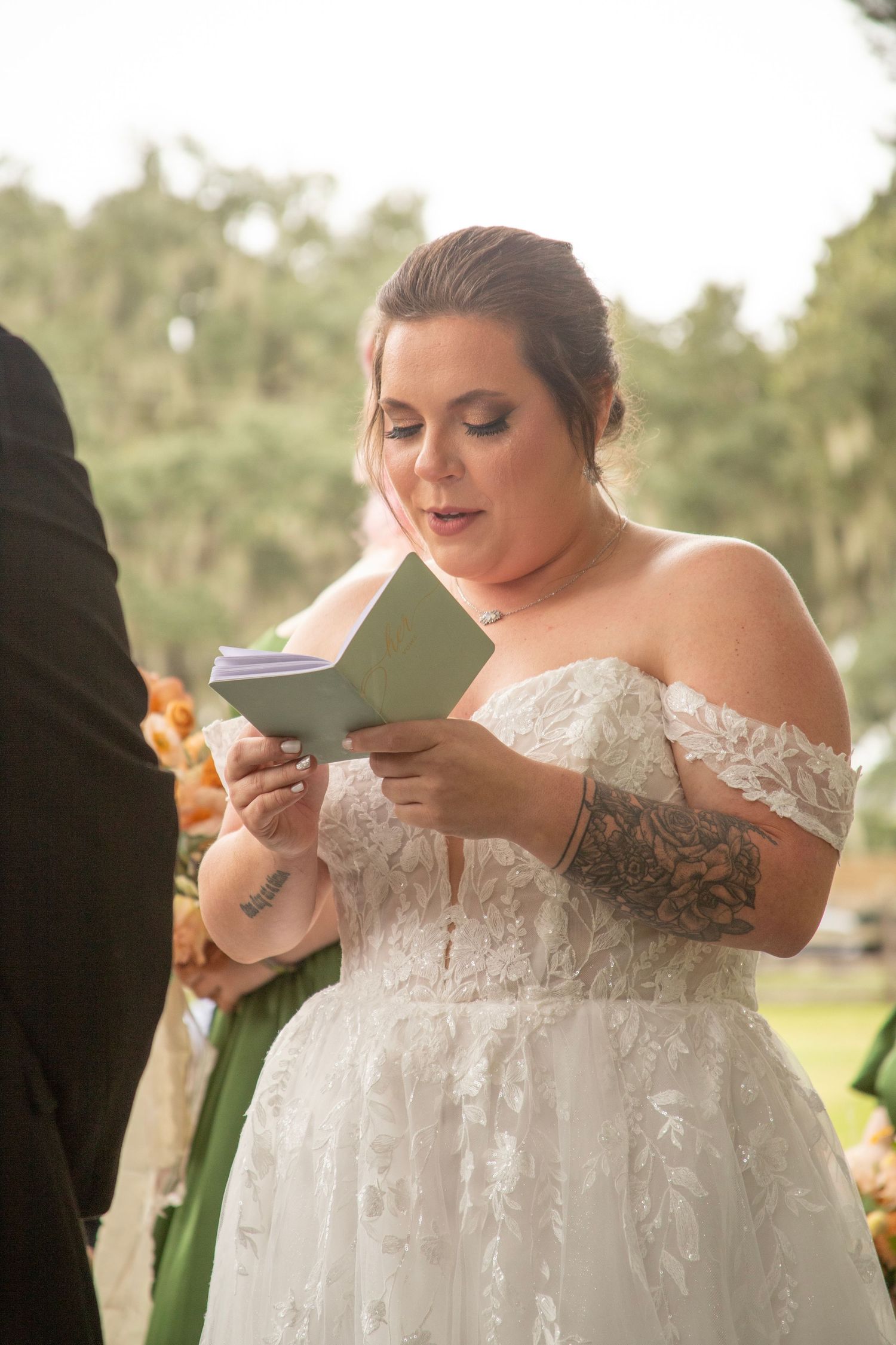 A bride in a white lace off-shoulder wedding dress reads from a paper during an outdoor ceremony.