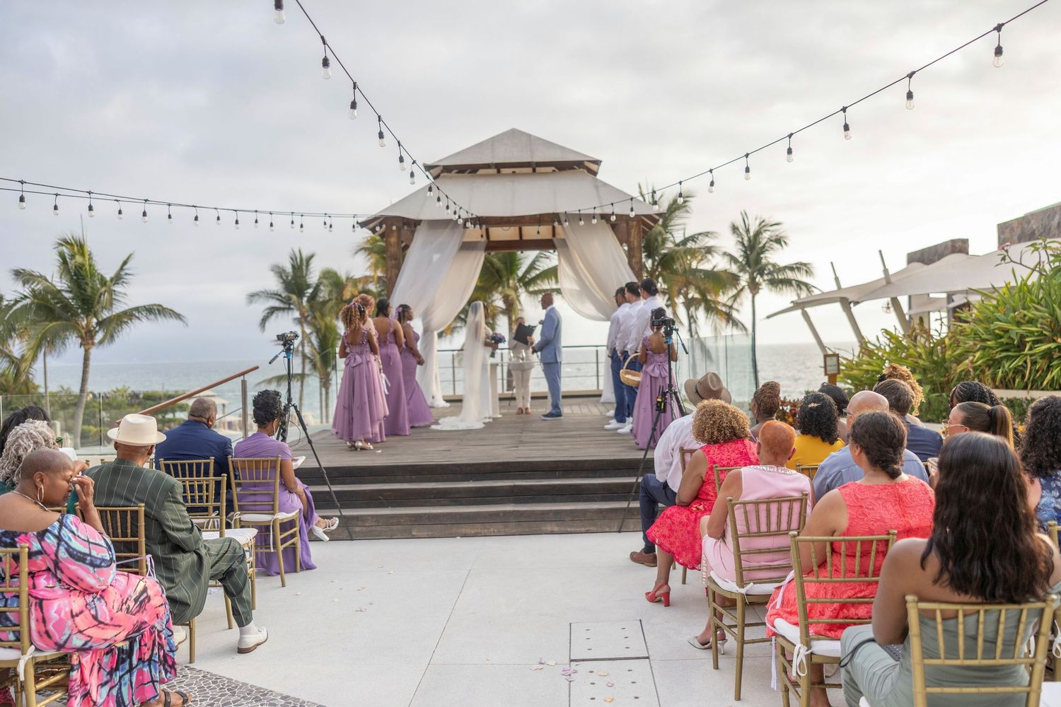 Outdoor wedding ceremony at a beachside gazebo with string lights and palm trees during a beautiful tropical sunset.
