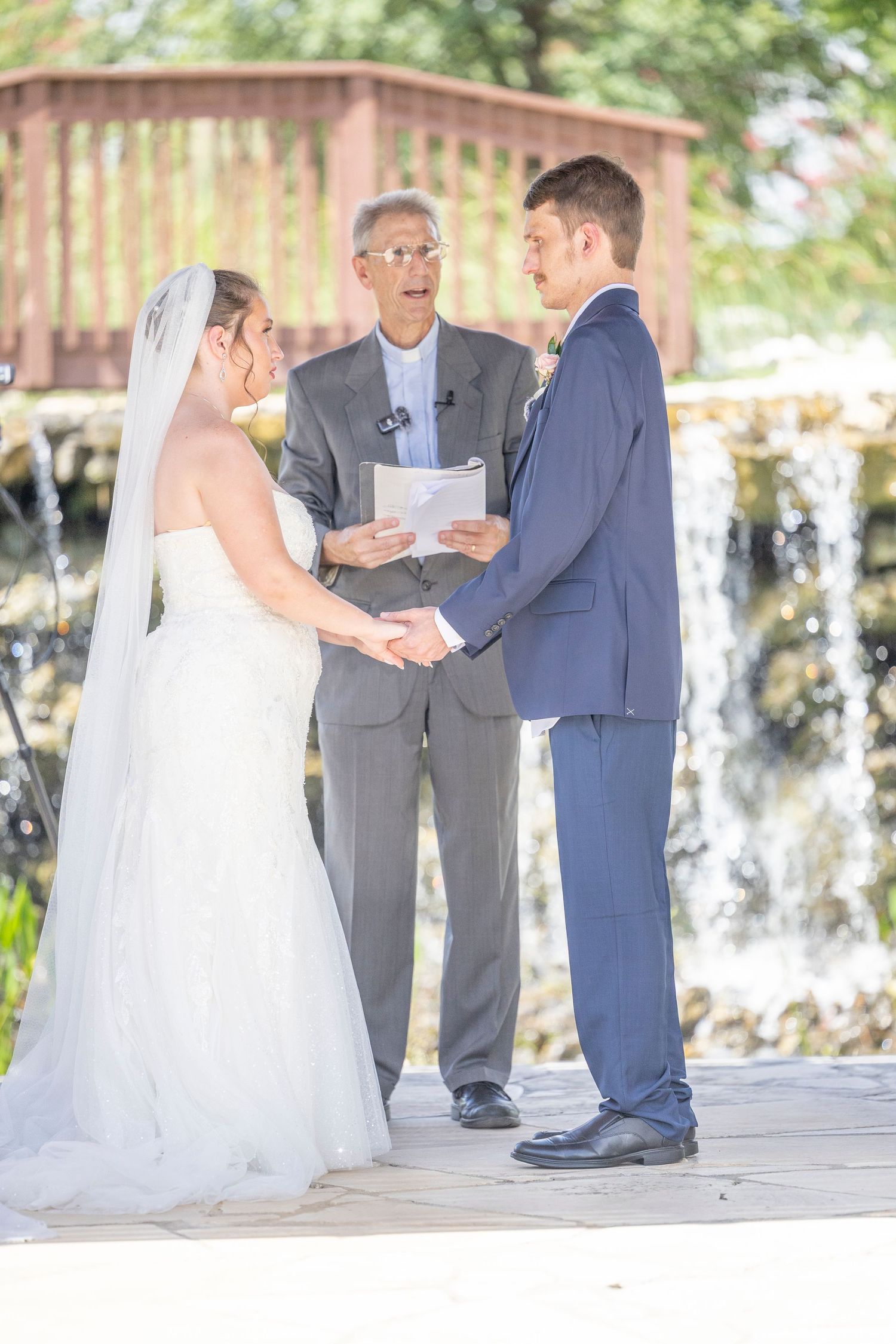 A wedding ceremony takes place outdoors with a waterfall backdrop as the couple exchanges vows with an officiant.
