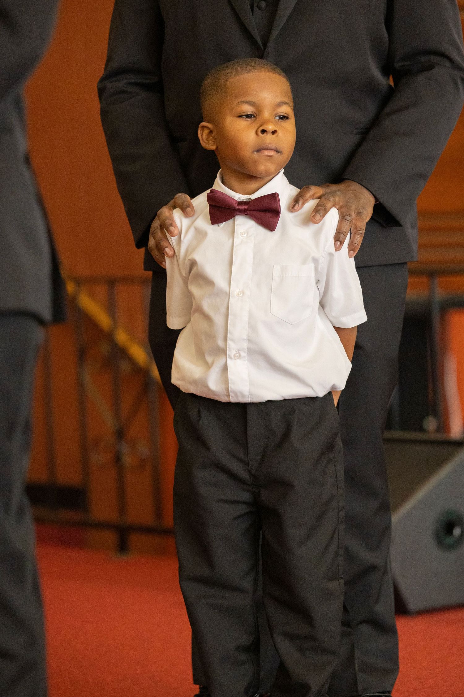 Young child in formal attire with white dress shirt, burgundy bow tie and black pants stands on red carpeted stage.