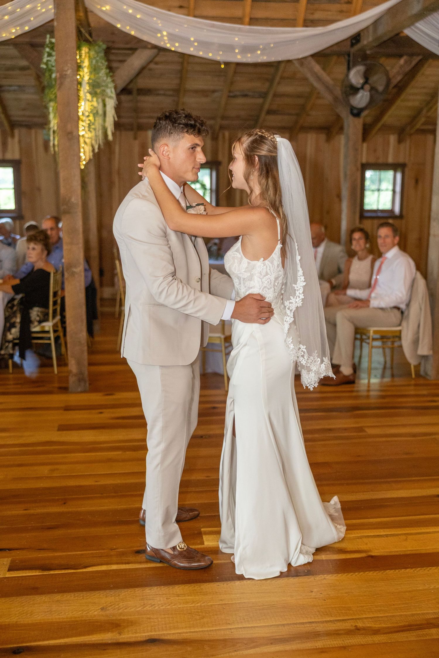 Newlyweds share their first dance on a polished wooden floor in a rustic barn venue decorated with string lights.