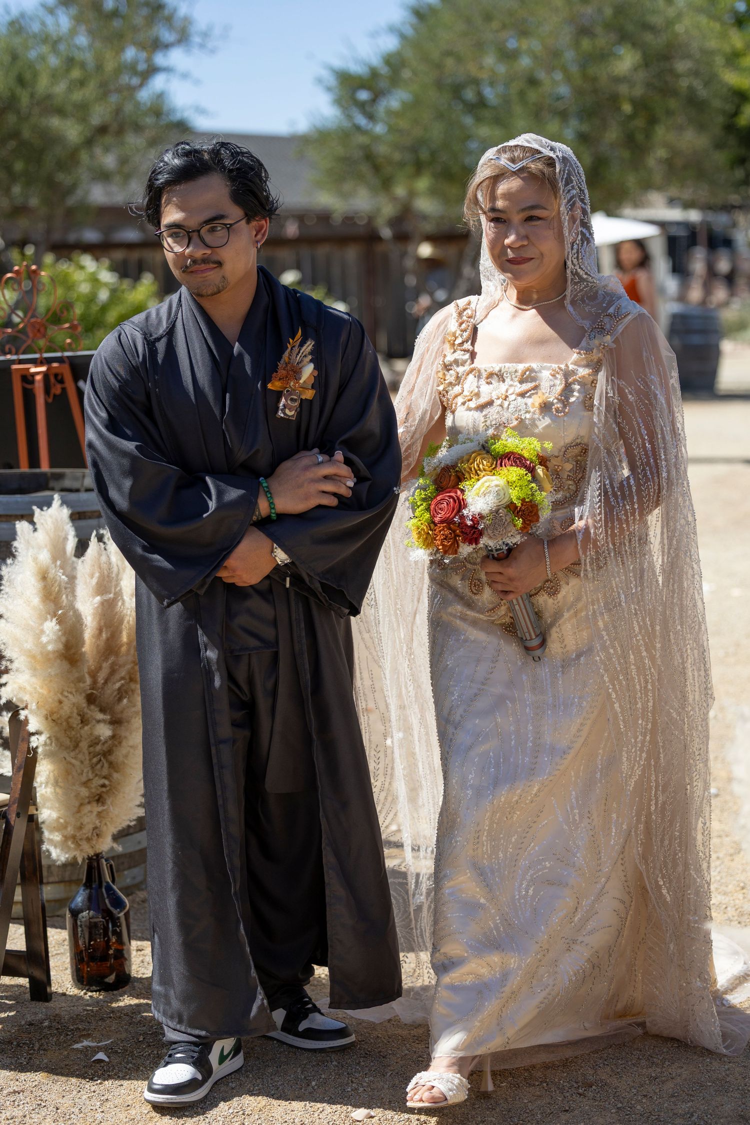 Bride in lace veil holding colorful bouquet stands beside groom in dark kimono at outdoor desert wedding ceremony.