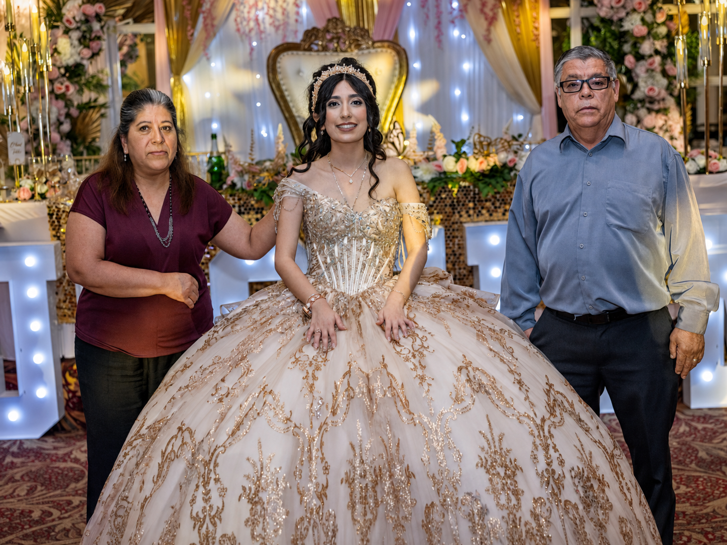 A young woman in a gold quinceañera ball gown poses with two adults at an elegant decorated celebration venue.