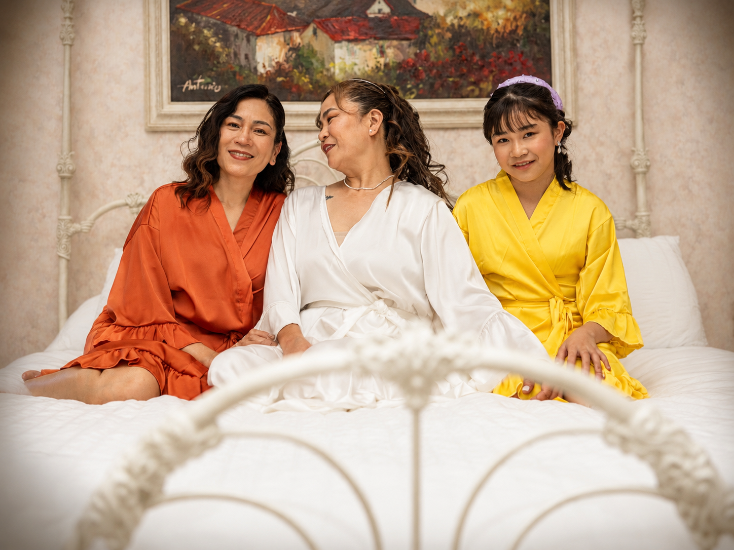 Three women in colorful satin robes sitting together on a white bed, smiling warmly in a cozy bedroom setting.