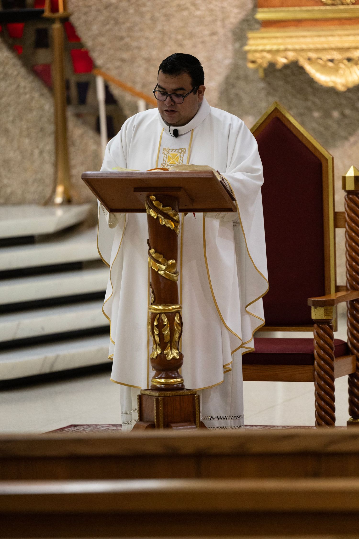 Catholic priest in white vestments reads from a golden lectern during a church Mass ceremony.