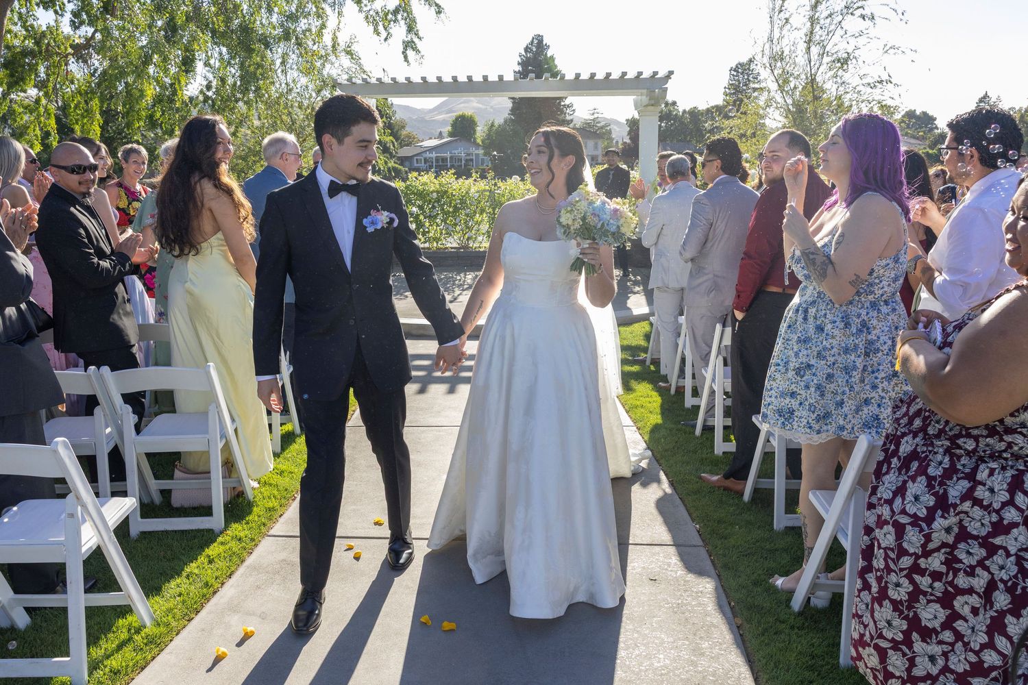 Bride and groom walk down the aisle hand in hand at an outdoor wedding ceremony with guests applauding.