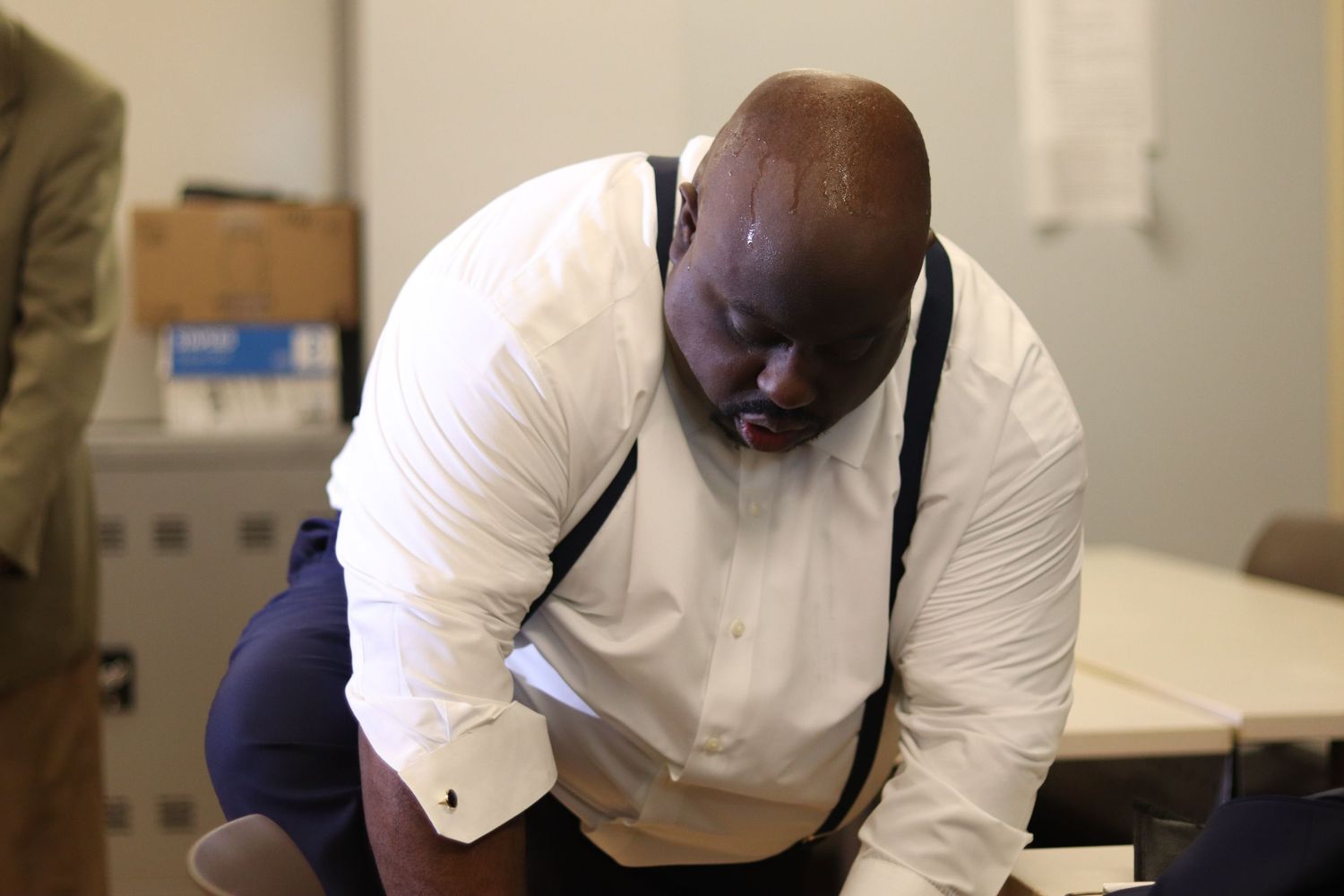 A large man in a white dress shirt and suspenders leans over a desk, reviewing documents in an office setting.