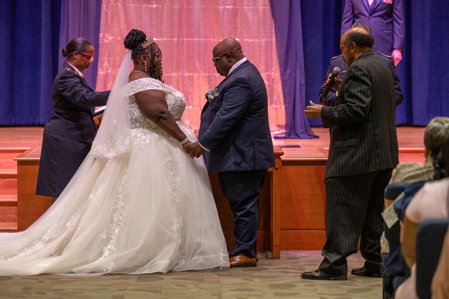 Bride and groom exchanging vows at wedding ceremony with officiant, bride in white gown, groom in navy suit.