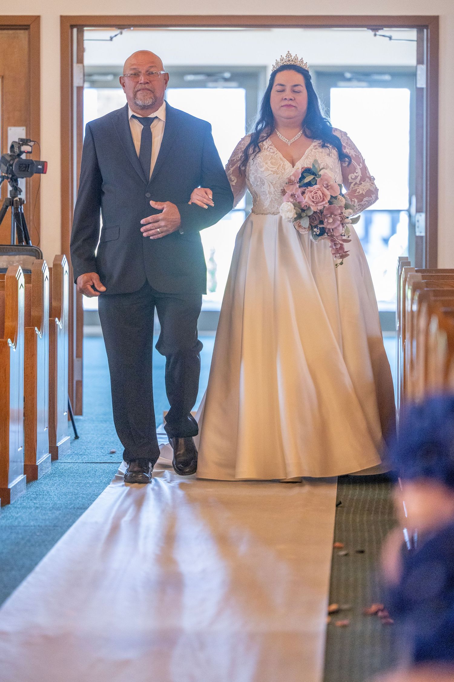 Bride in white gown with bouquet walks down church aisle with escort during traditional wedding ceremony.