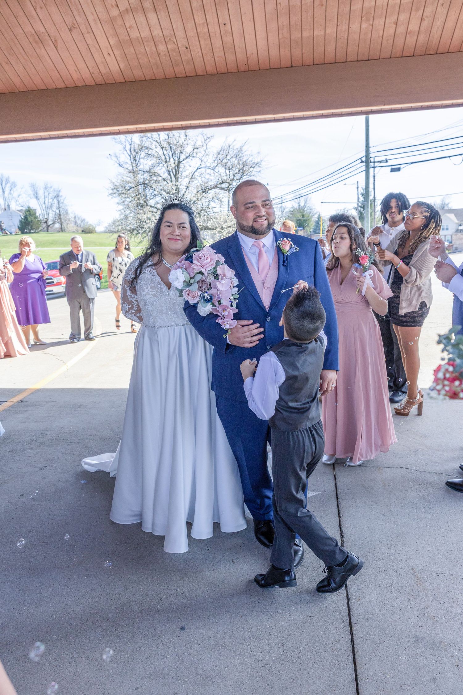 Bride in white gown and groom in blue suit exit wedding ceremony with guests celebrating and child hugging groom.