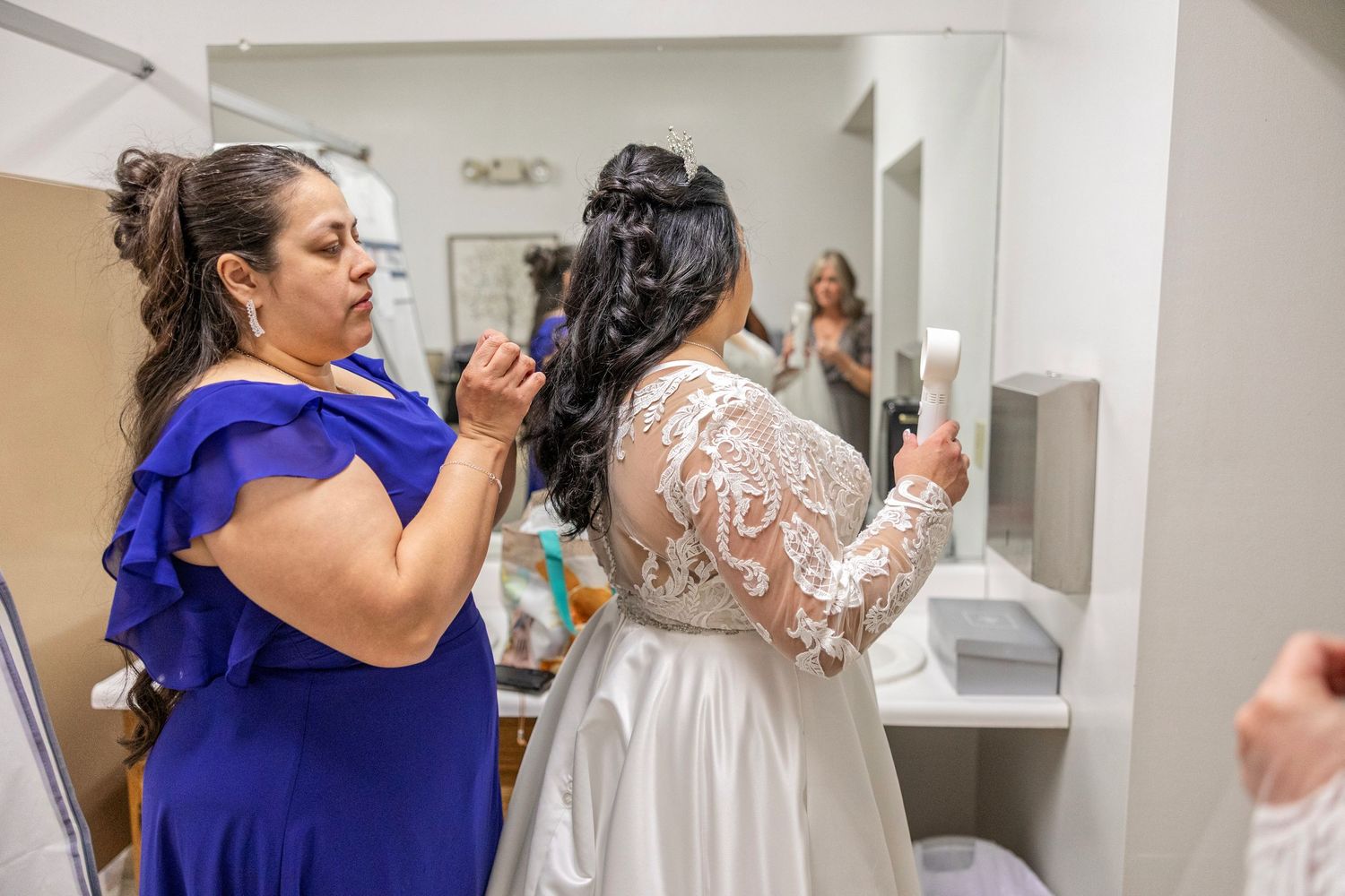 Bride in lace wedding gown gets hair styled by woman in blue dress in a bridal preparation room.