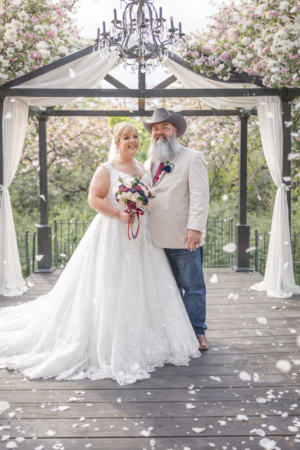 Bride in white gown holding bouquet stands with bearded groom in cowboy hat under floral gazebo with falling petals.