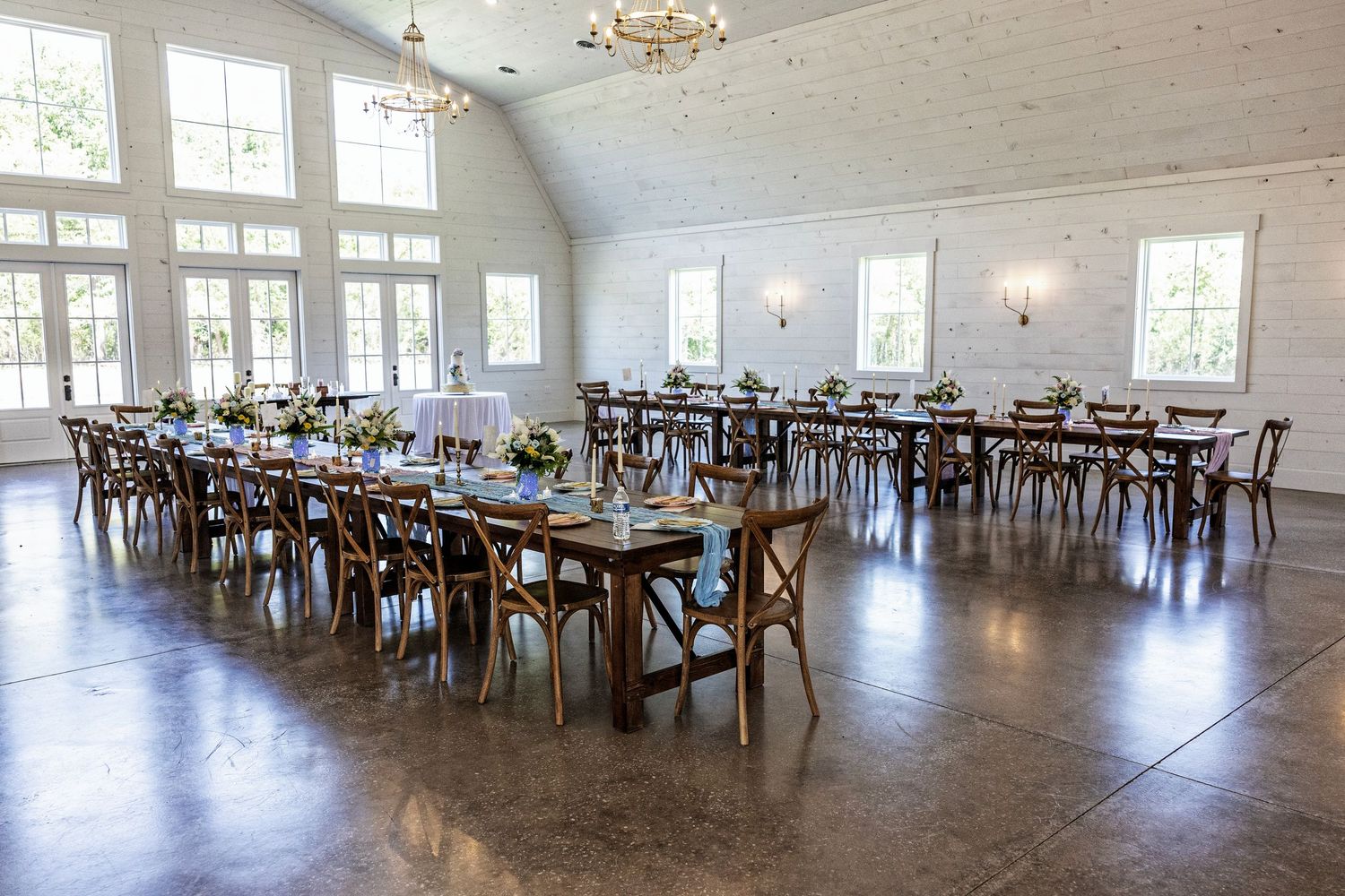 Elegant wedding reception hall with rustic wood farm tables, cross-back chairs, floral centerpieces, and vaulted white ceilings.