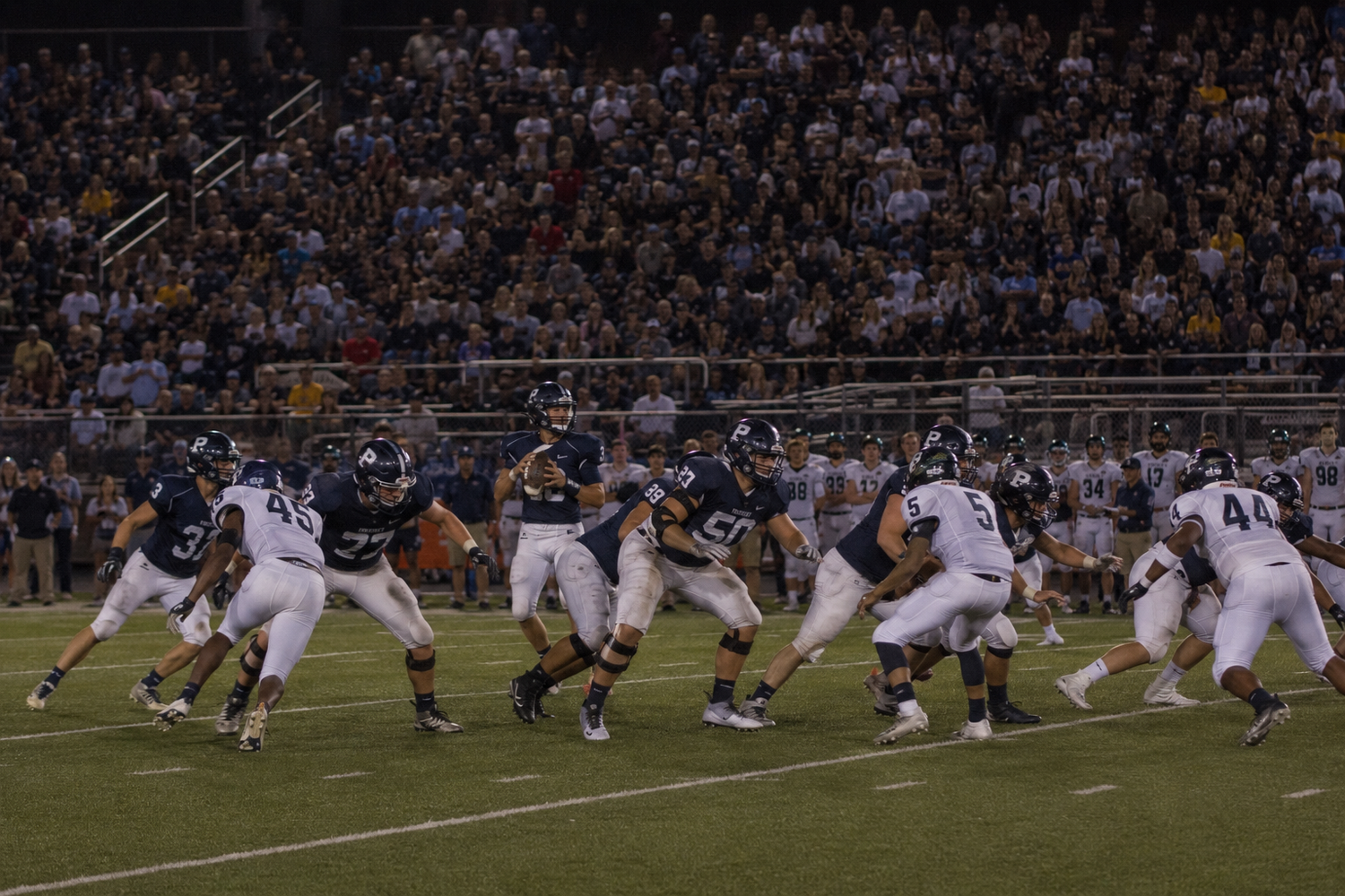 Football players clash at the line of scrimmage during an intense night game before a packed stadium crowd.