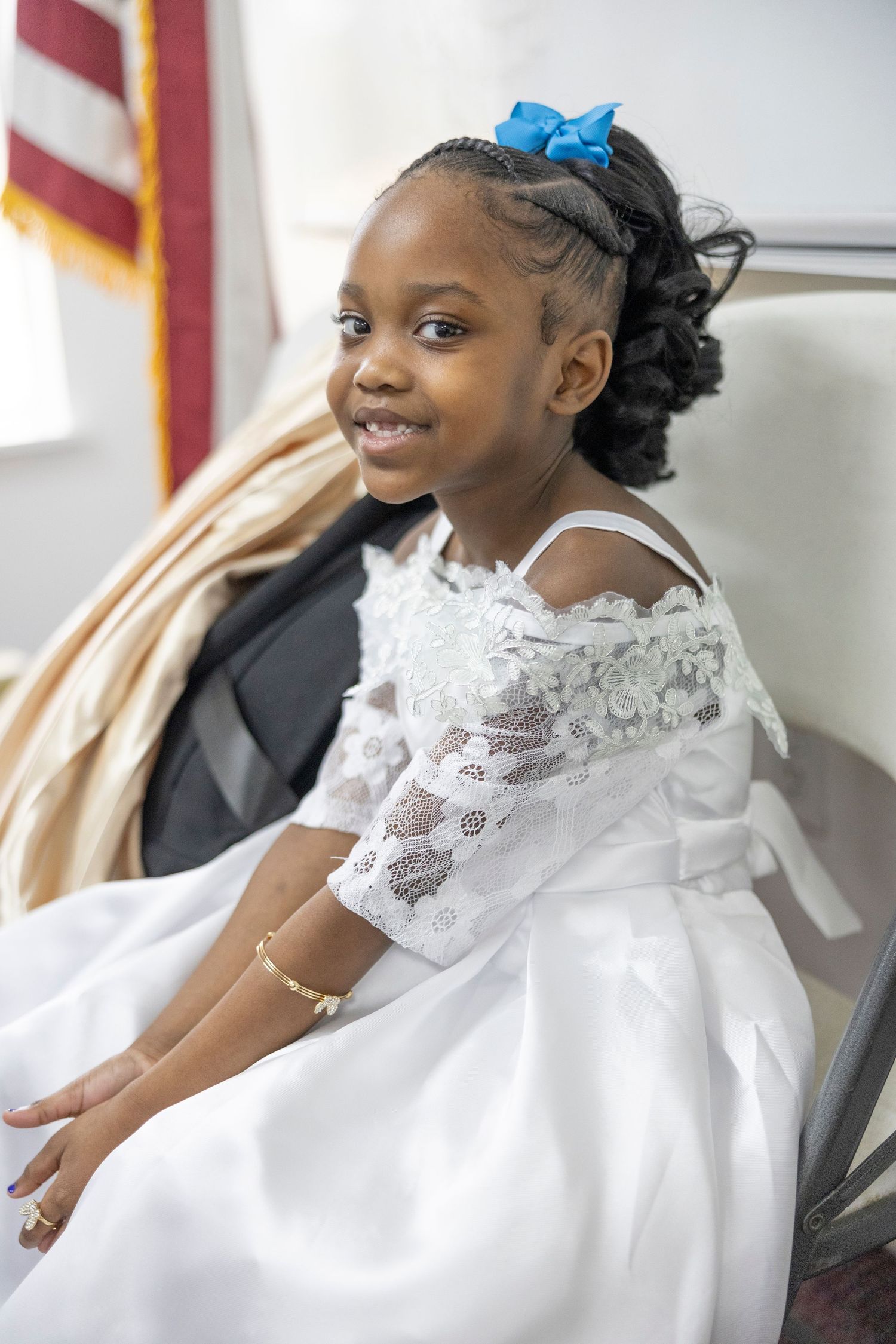Young girl in white lace dress with blue hair bow smiles while seated, wearing gold bracelet.