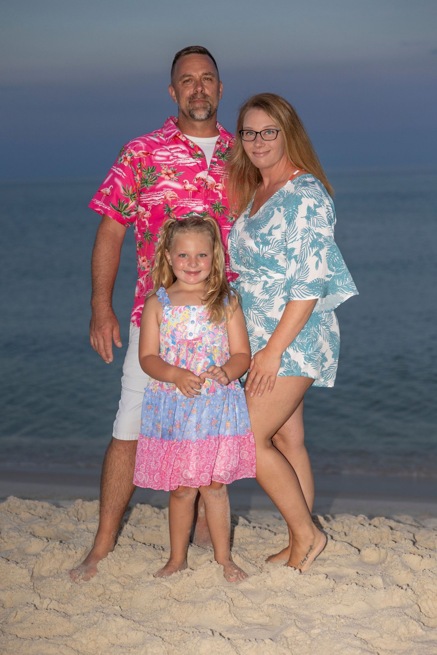 A family of three wearing coordinated tropical prints poses together on a beach at dusk.