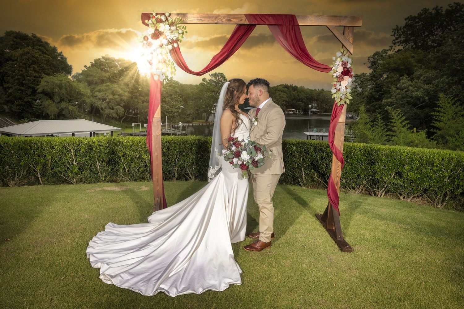 Wedding couple embraces under draped red arch during golden sunset on manicured lawn.