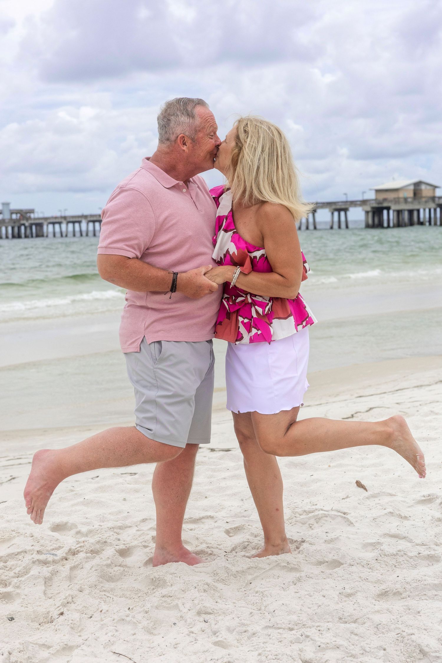 Playful couple in pink and white outfits having fun on the beach with ocean pier in background.