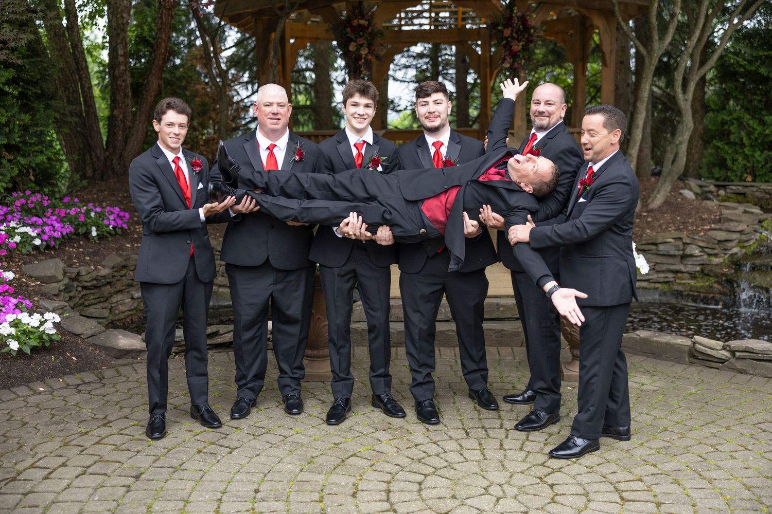 Wedding party in dark suits and red ties share a candid moment while standing together outdoors.