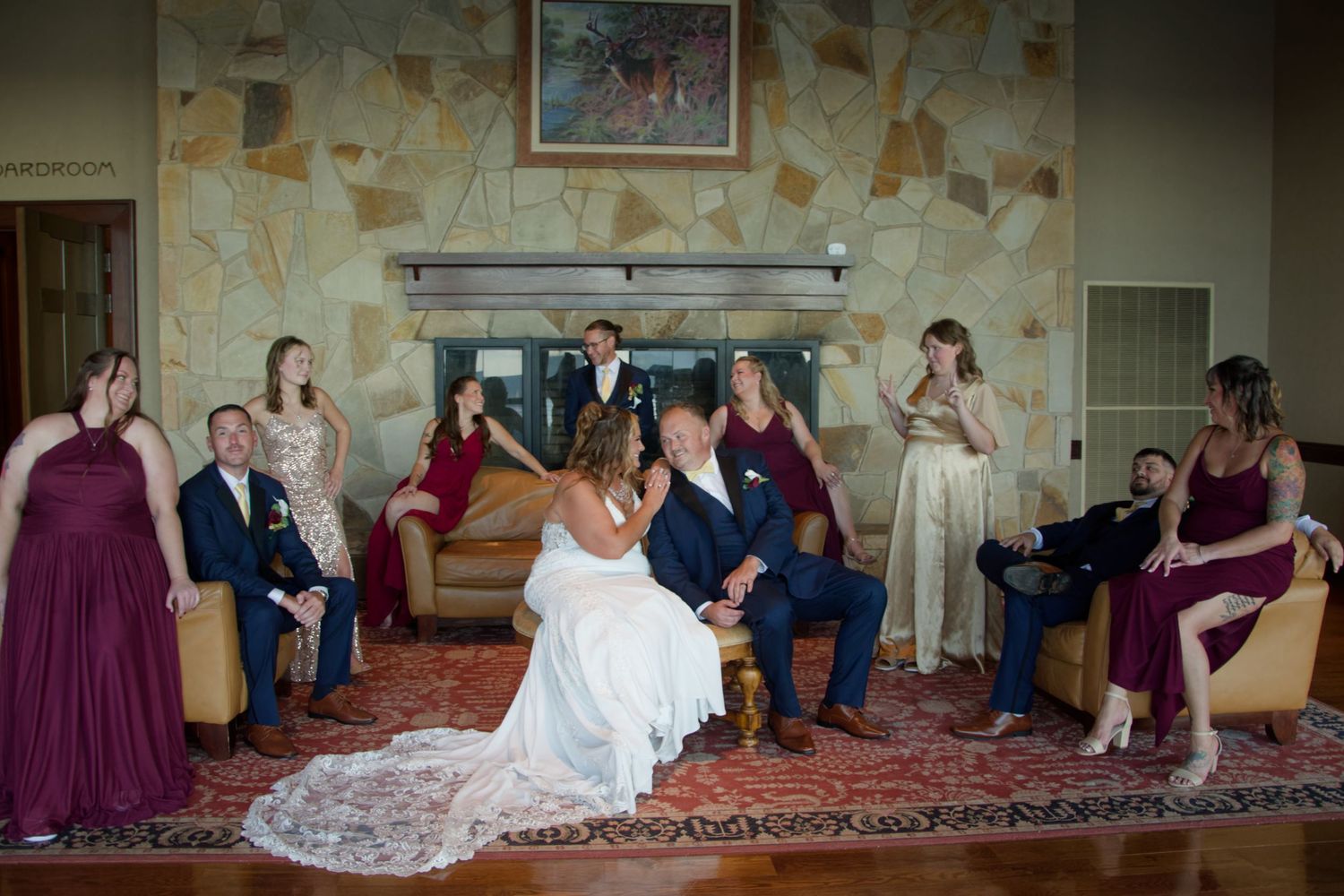 Wedding party members in burgundy dresses and formal attire gather for a fun group photo session on a carpeted floor.