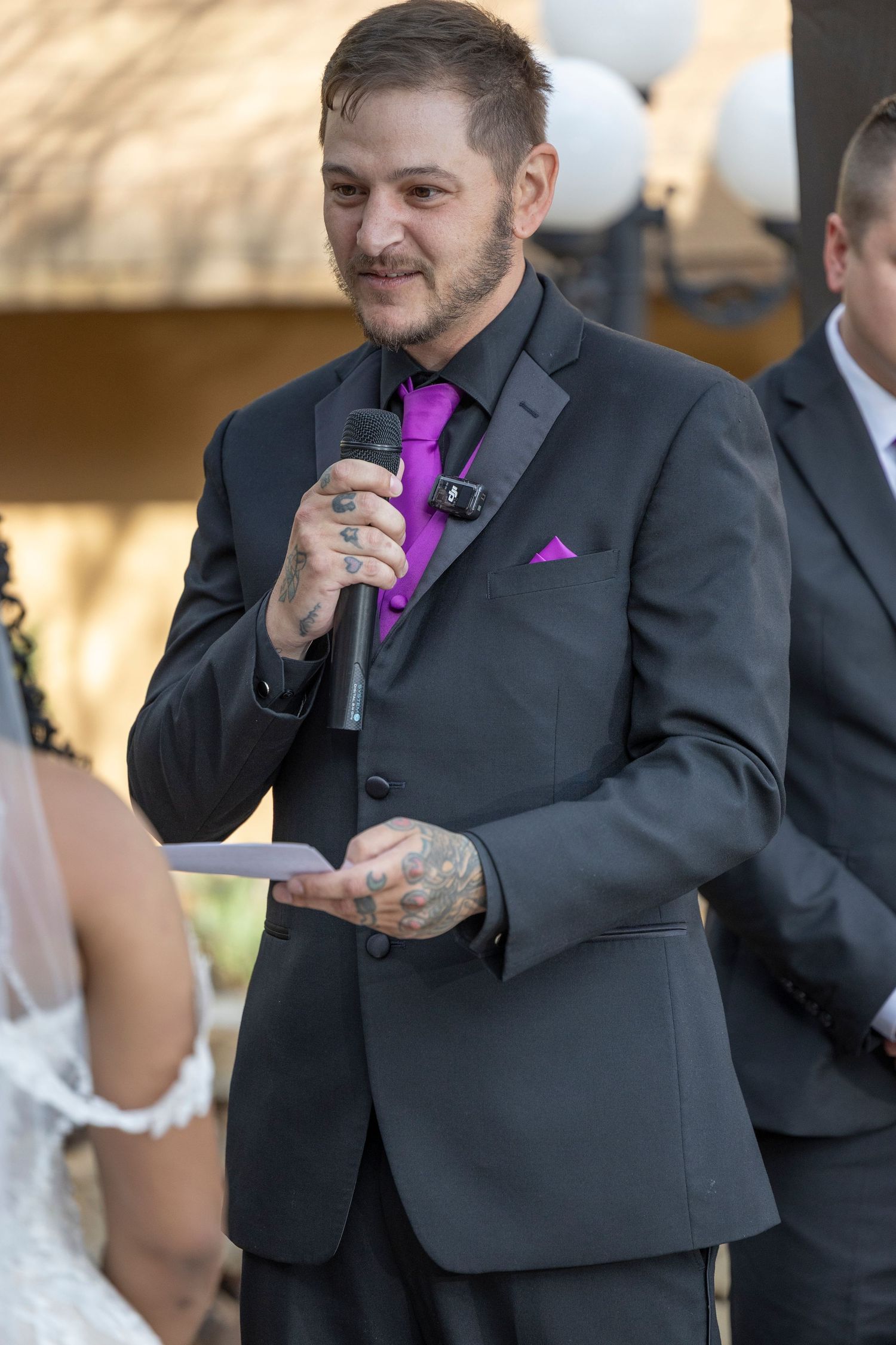 A person in a dark suit with a purple pocket square speaks into a microphone during a formal event.